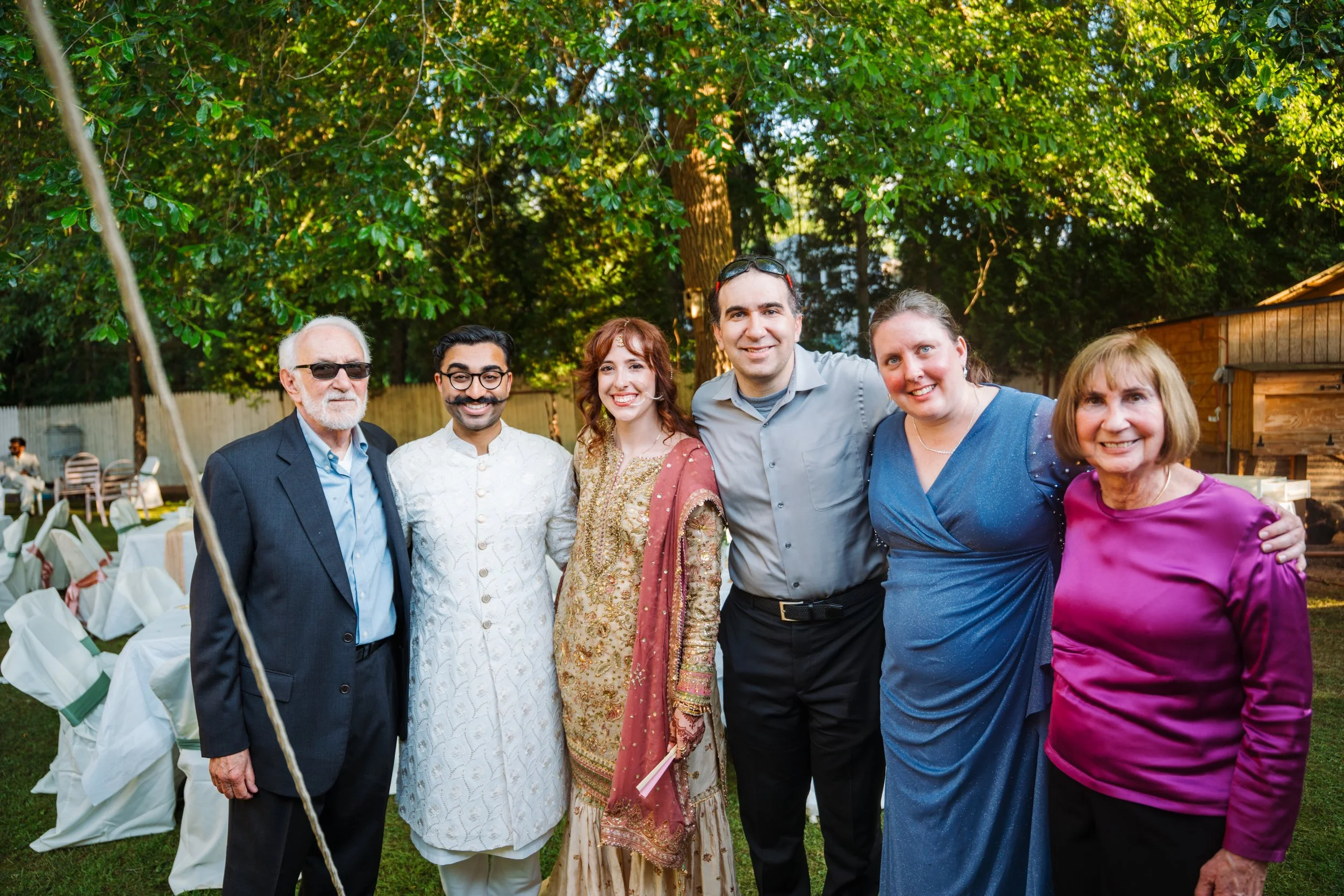 Group of six diverse people standing outdoors at a celebration, smiling, with trees and tables with white chairs in the background.