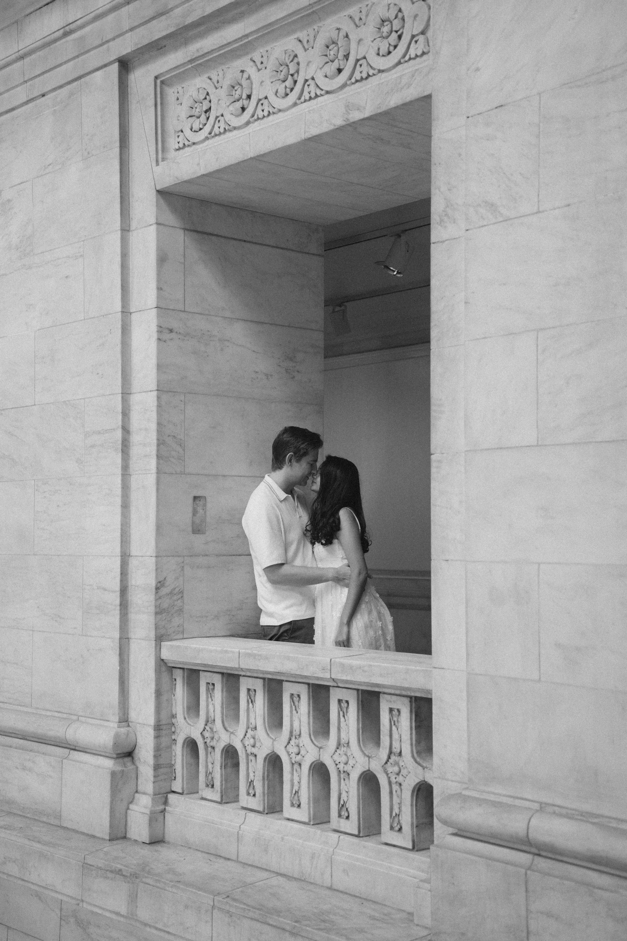 A young couple is standing close together on a stone balcony in front of a large opening in a marble wall, with ornate carved details, possibly at a historic or government building.