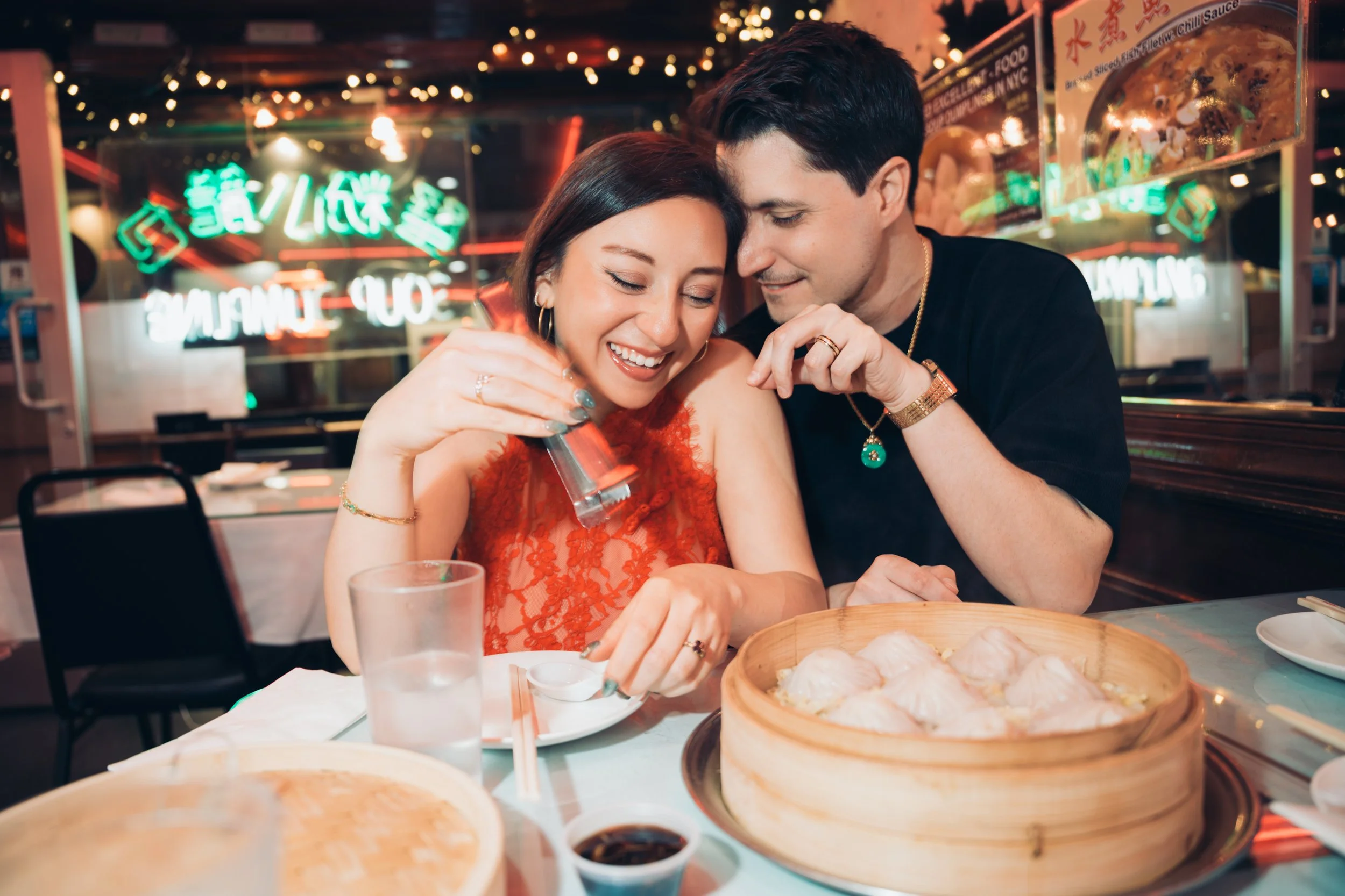 A couple enjoying a meal at a restaurant with neon lights in the background. They are smiling and looking at each other, surrounded by sushi and drinks on the table.