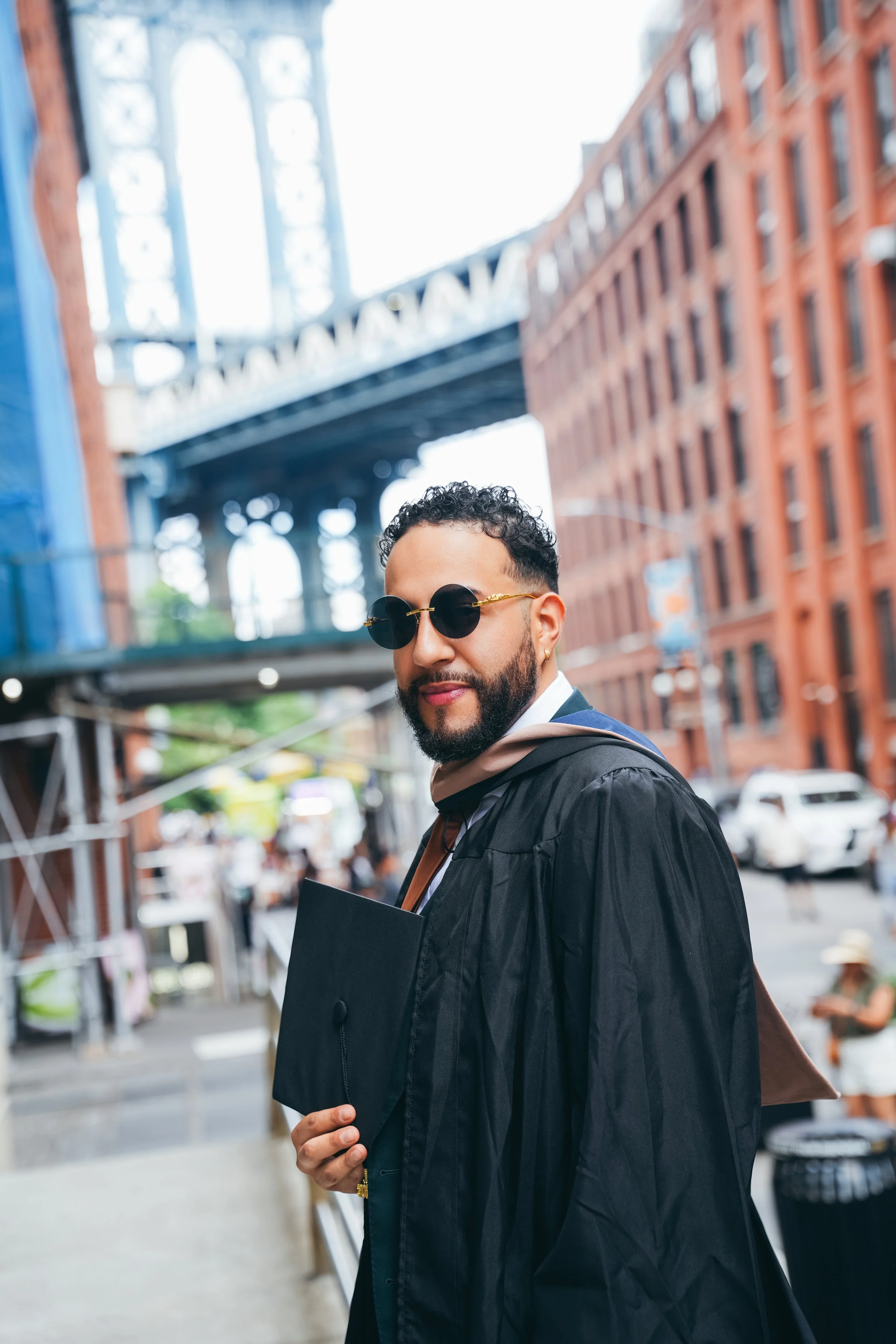 A man with curly hair and sunglasses dressed in a graduation gown, holding a folder, standing on a city street with red brick buildings and a bridge in the background.