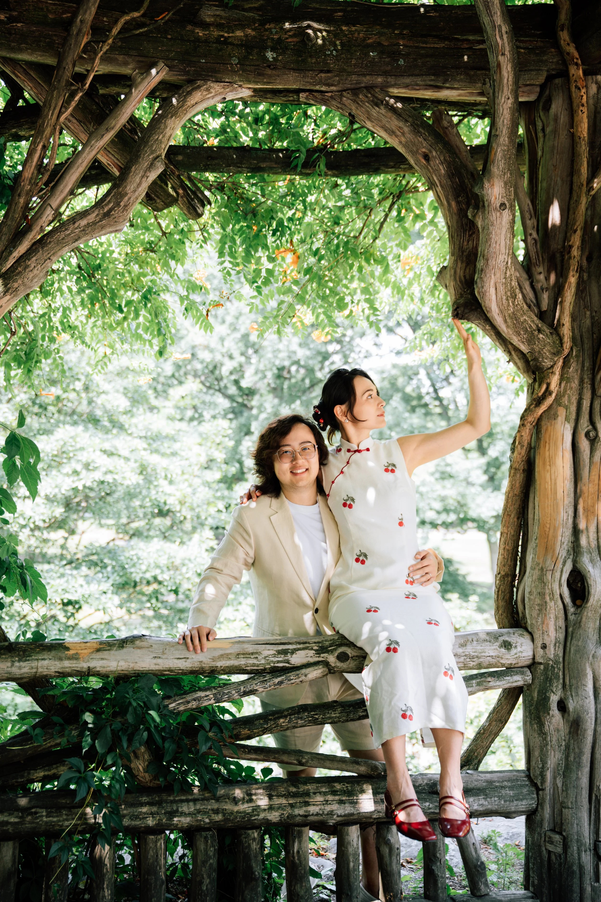 Two women are sitting on a wooden log within a tree house surrounded by green foliage. One woman, with dark hair in a bun and dressed in a traditional cream-colored dress with red cherry patterns, is looking up and is wearing red shoes. The other wom