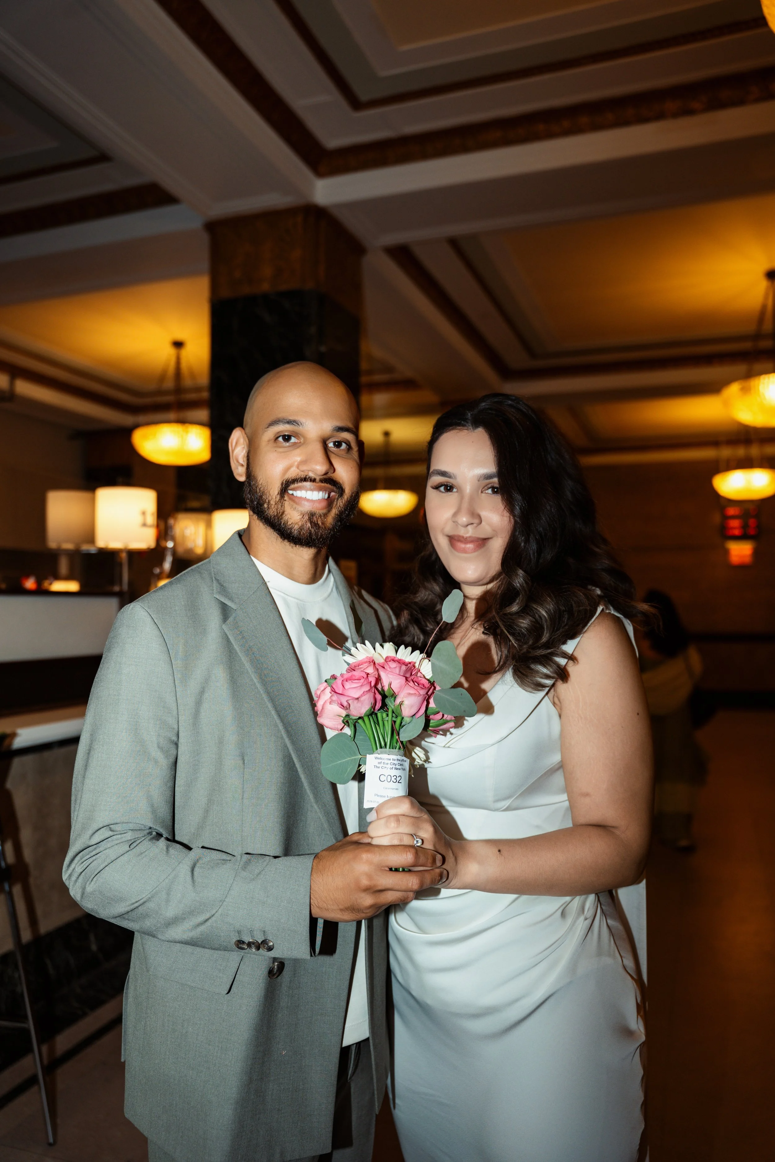 A man and woman at a formal indoor event, smiling and holding a bouquet of pink flowers together, with warm lighting and decorative ceiling in background.