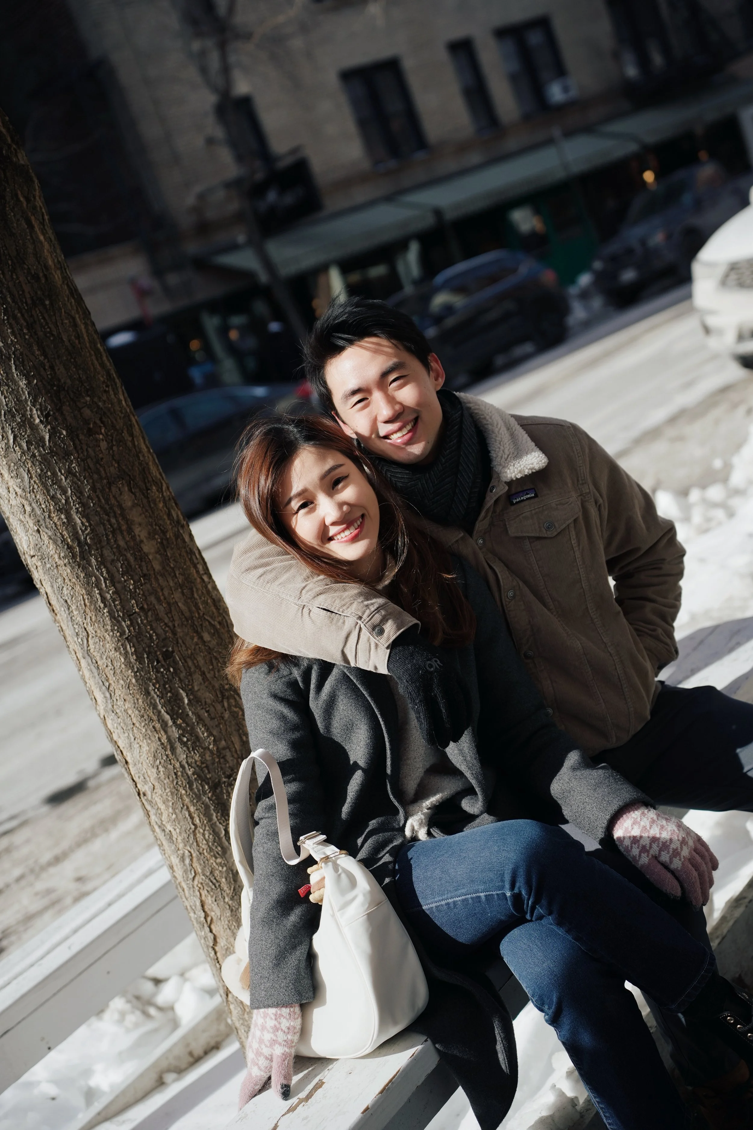 A smiling young man and woman sitting on a bench outdoors in winter, leaning against a tree, with snow on the ground and parked cars and buildings in the background.