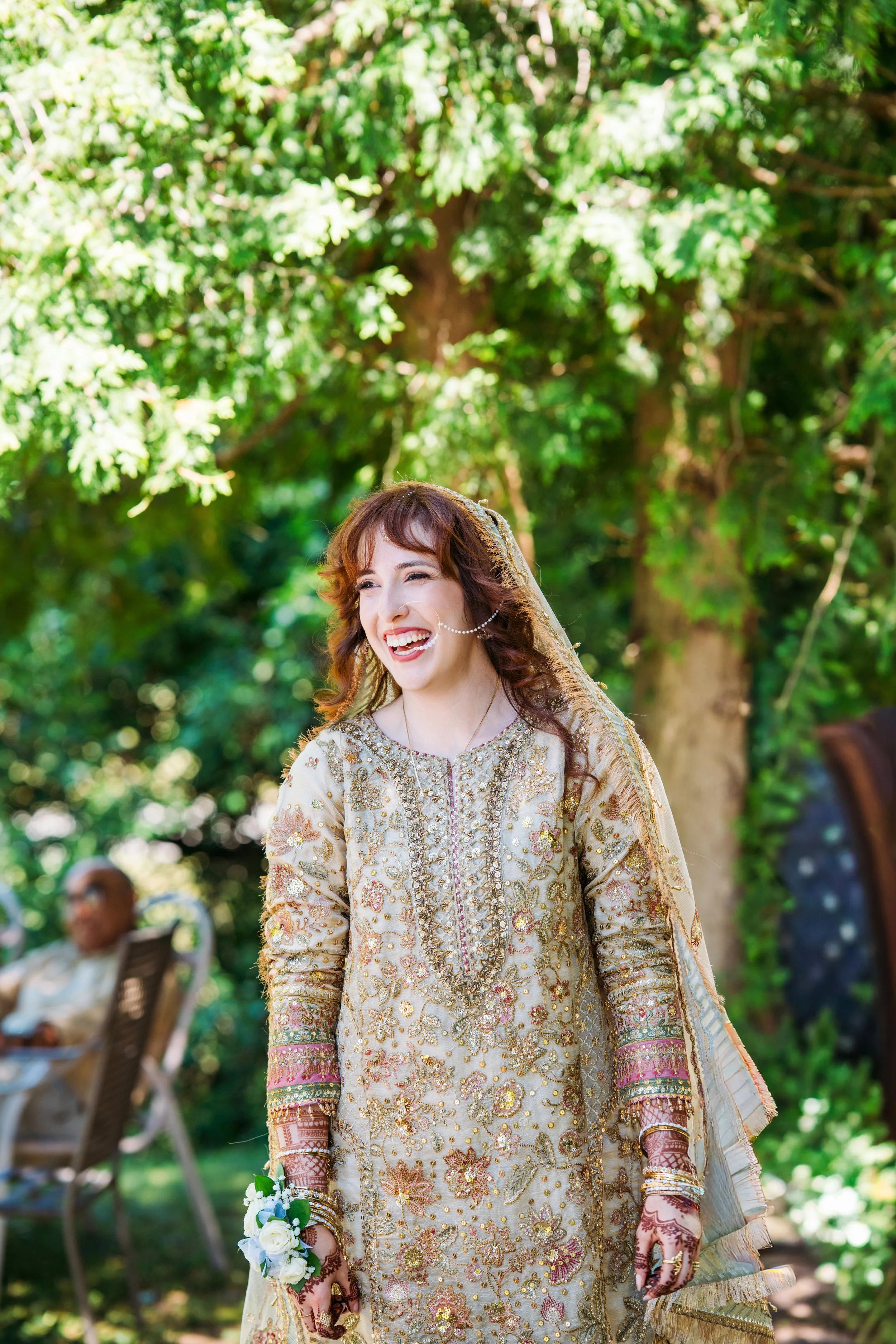 A woman in a traditional, ornate cream and pink dress with gold embroidery, holding a bouquet of white roses, standing outdoors under a green leafy tree, smiling.