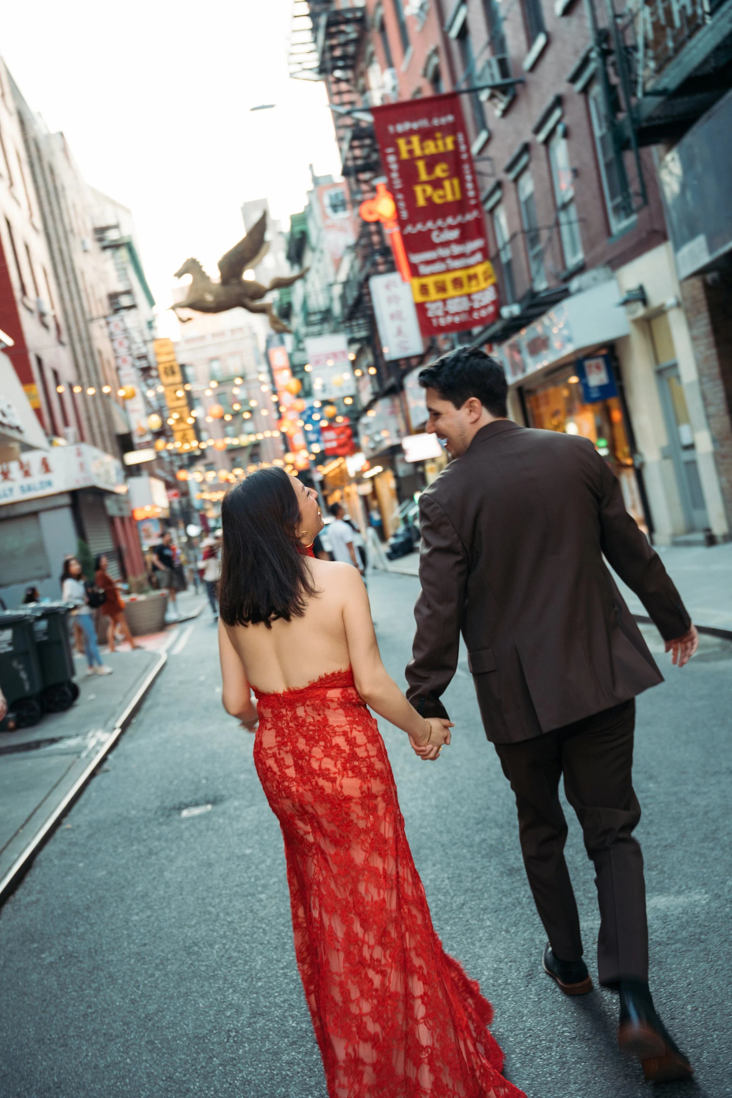 A man and woman walking hand-in-hand down a busy city street, smiling at each other. The woman is dressed in a red lace dress, and the man is wearing a dark suit. The street is adorned with hanging lights, and there are storefronts and signs, includi