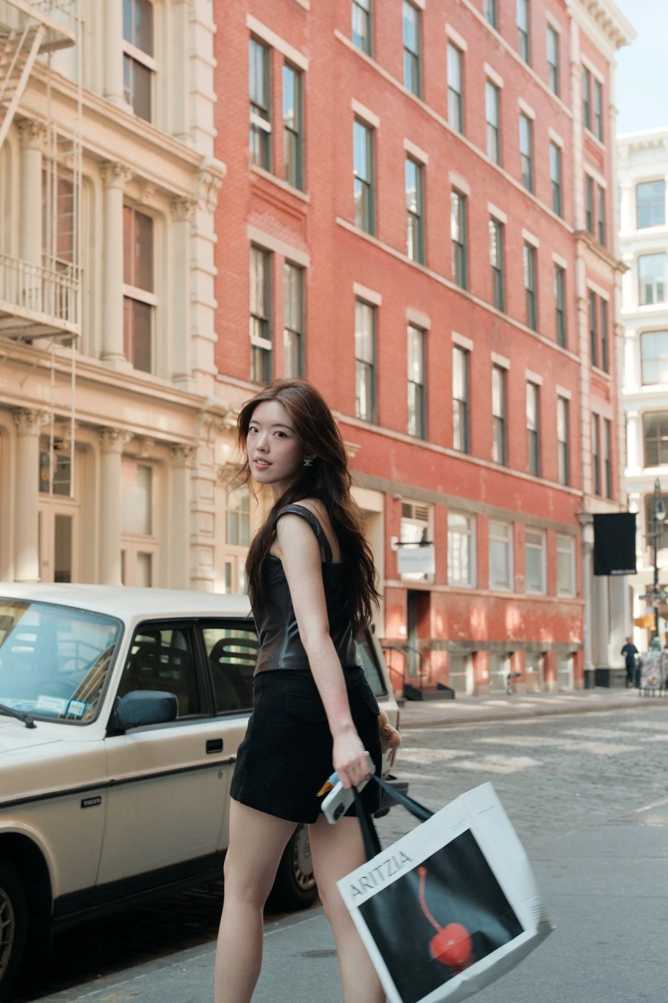A woman with long dark hair wearing a black dress, holding a shopping bag and a small object, standing on a city street beside a vintage beige car with a European license plate, with old brick buildings in the background.