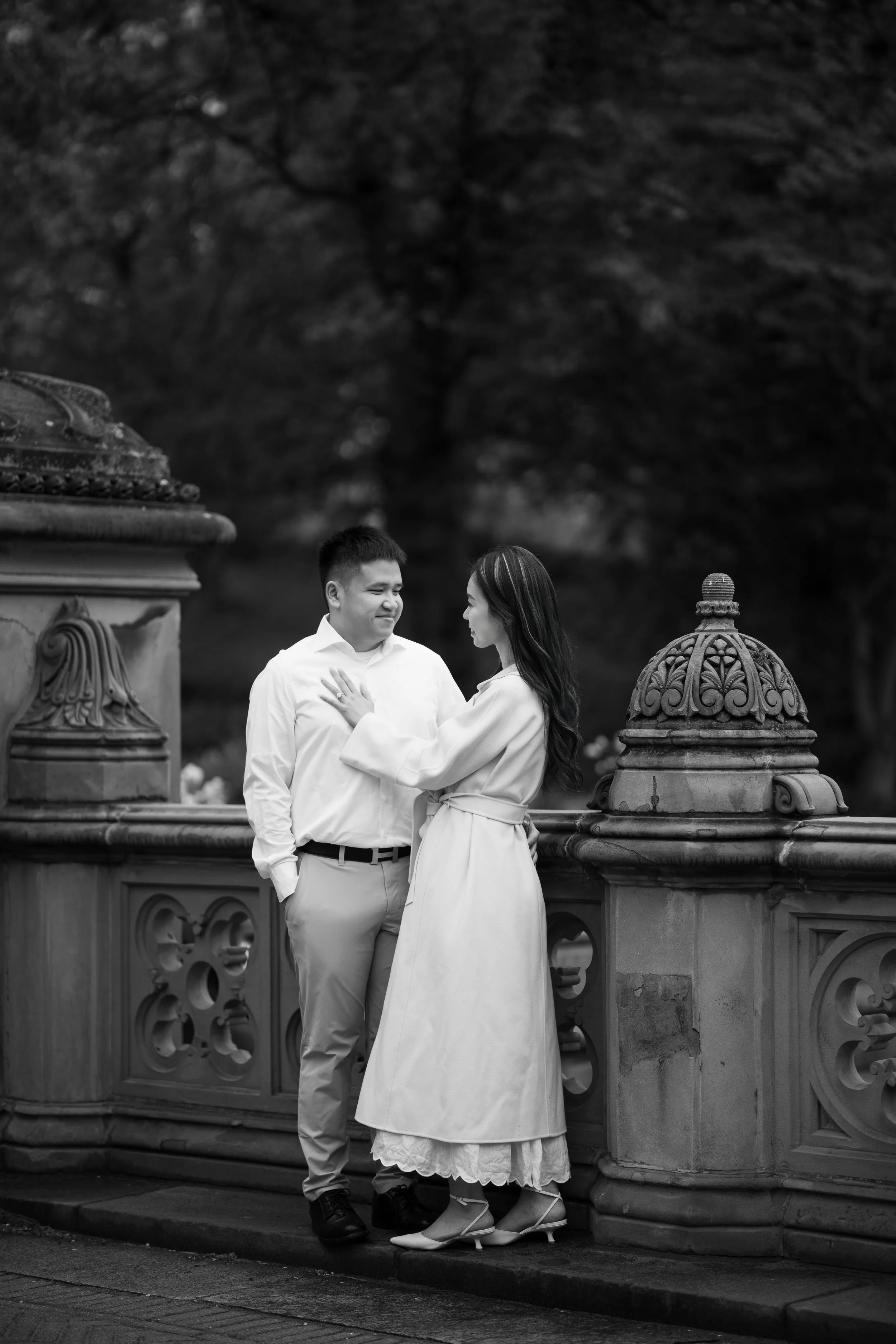 A couple dressed in white standing close on a decorative stone bridge with ornate railings, surrounded by trees, with the woman gazing at the man, in a black-and-white photograph.