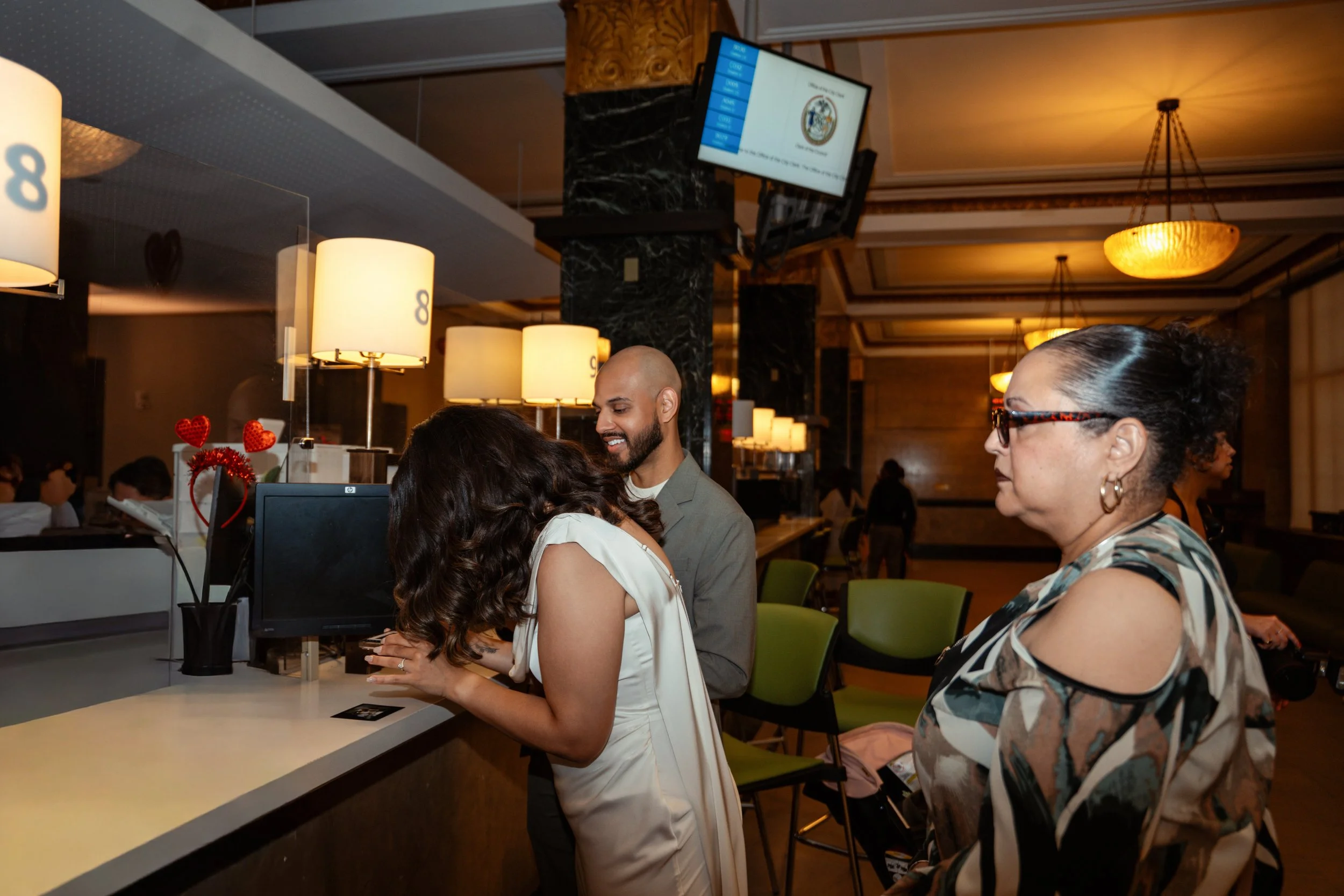 A woman in a white dress and a woman with glasses and a patterned top standing in line at a hotel reception desk, talking to a hotel employee with a beard and a gray blazer. The reception area has warm lighting, lamps, a computer, and decorations wit