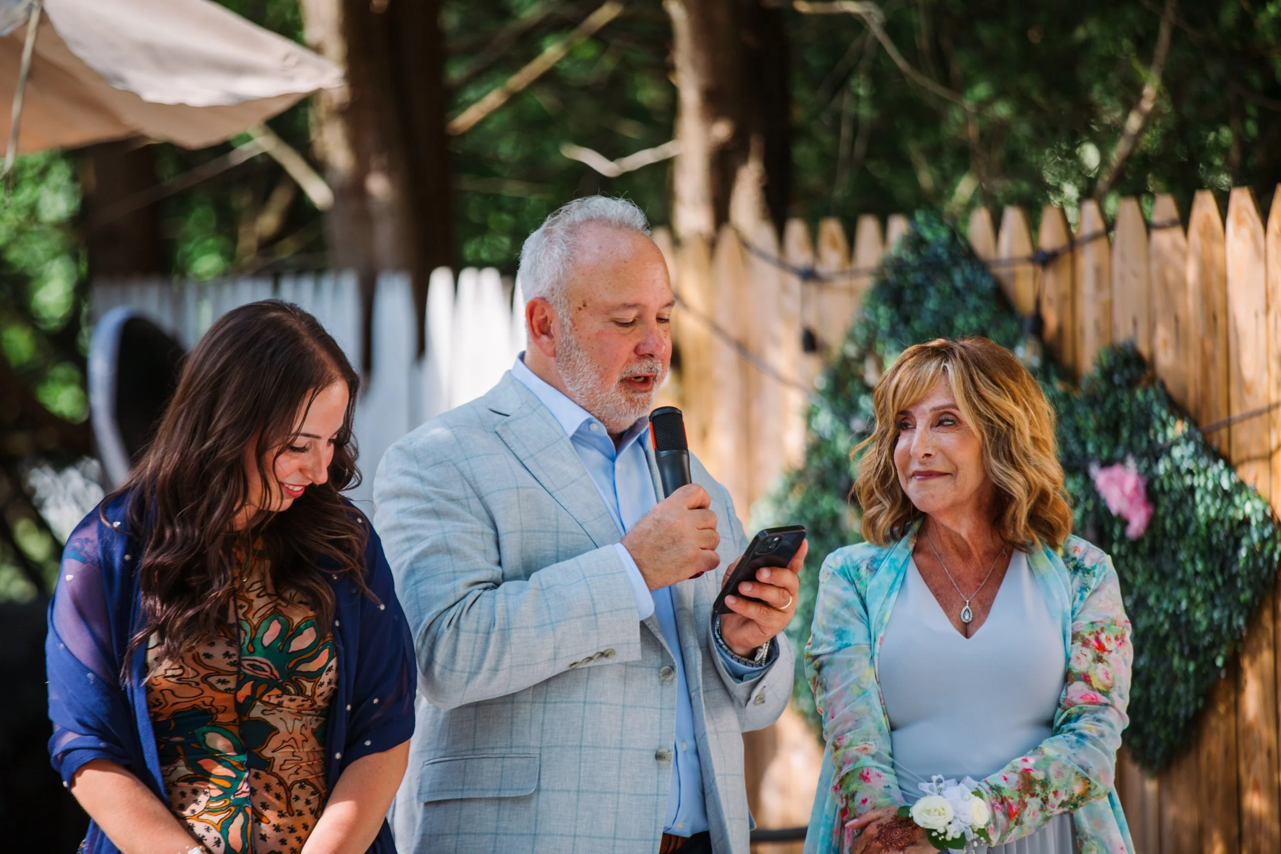 An elderly man in a light gray suit is speaking into a microphone while holding a phone, standing between two women during an outdoor event. One woman on his right is smiling and has long dark hair, dressed in colorful attire. The woman on his left h