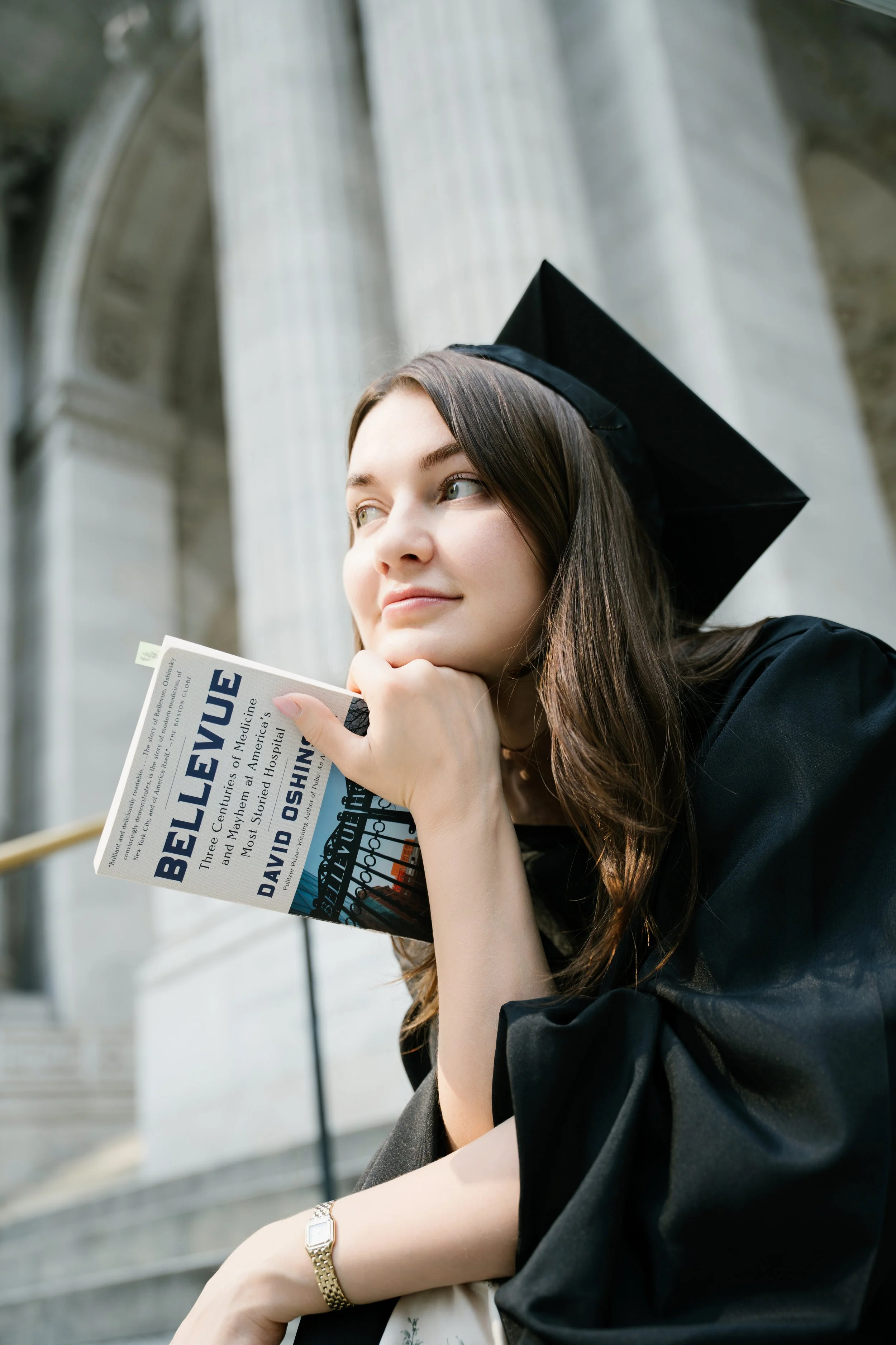 A young woman in a black graduation cap and gown reads a newspaper outside a building with large white columns.
