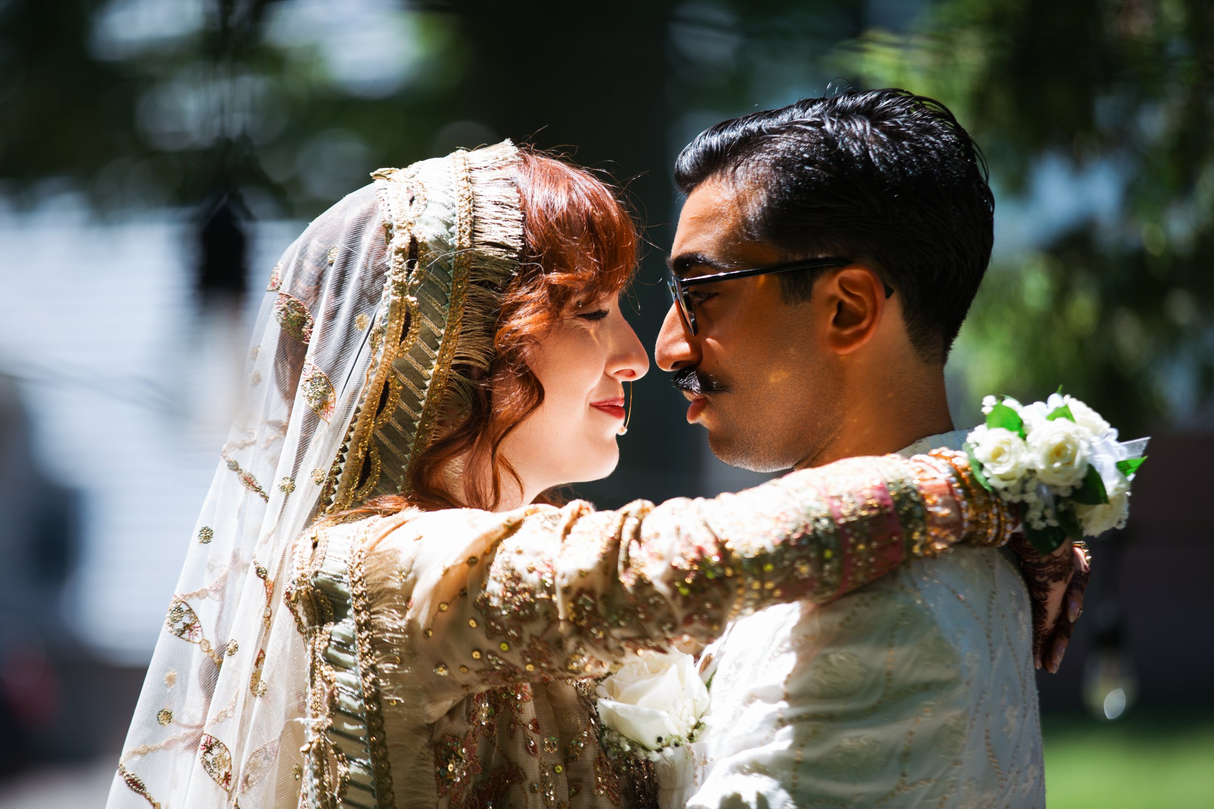 A couple in traditional Indian wedding attire touching foreheads, outdoors with sunlight and blurred greenery in the background.