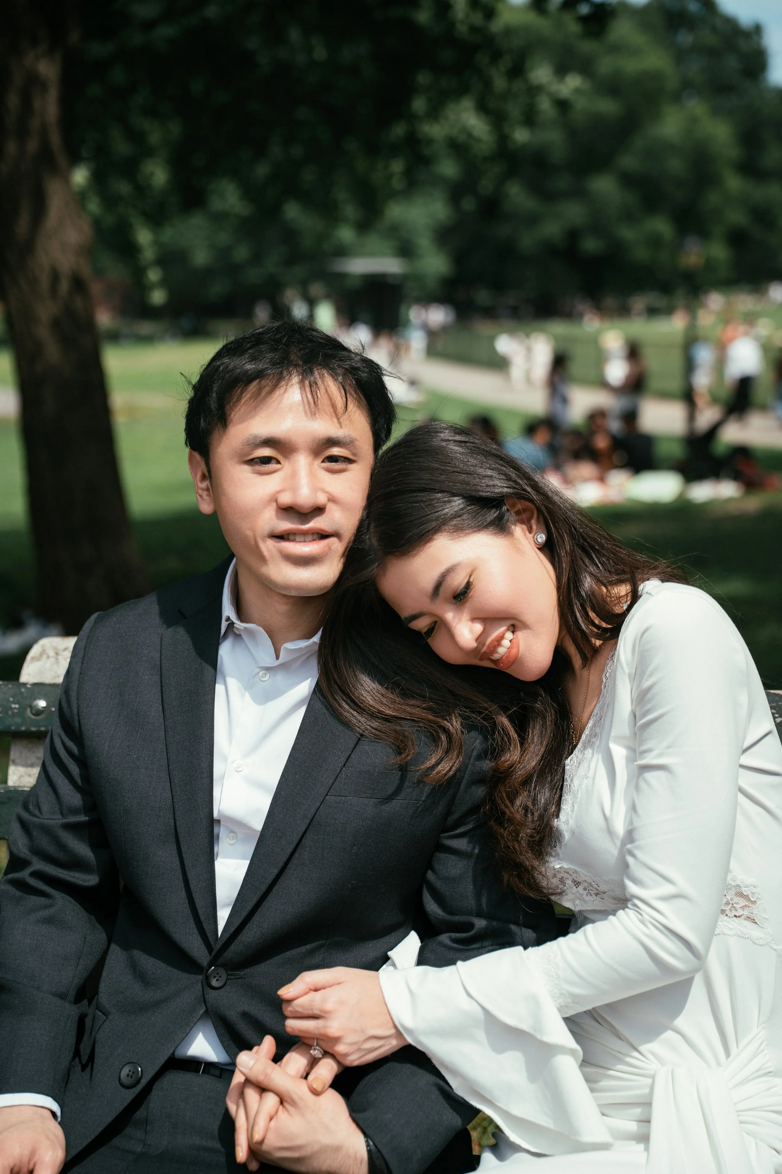 A man in a gray suit and a woman in a white dress are sitting on a park bench, hugging and smiling. The background shows a park with trees and other people enjoying the outdoors.