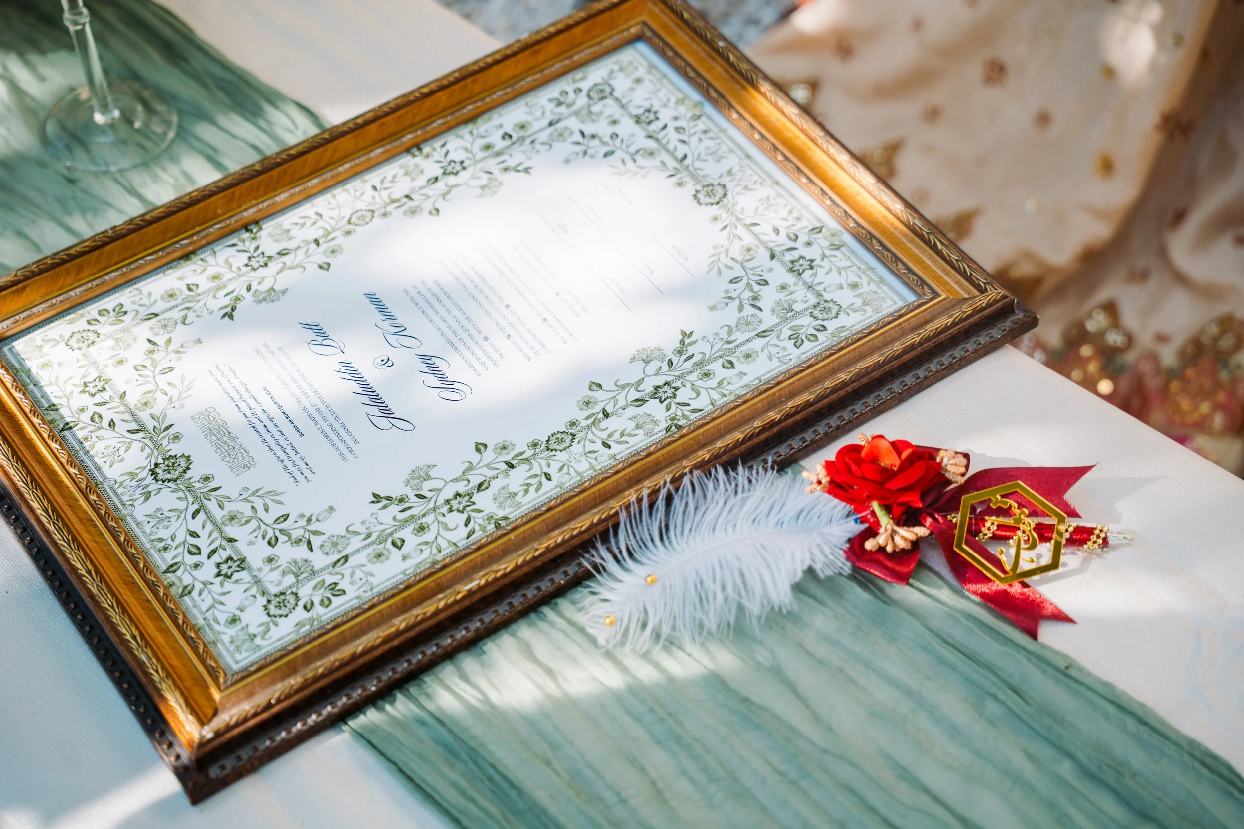 Decorative framed certificate placed on a table with a wine glass and a red ribbon tassel. The certificate has floral patterns and is awarded to Madison Blue and Jinger T. Hawn.