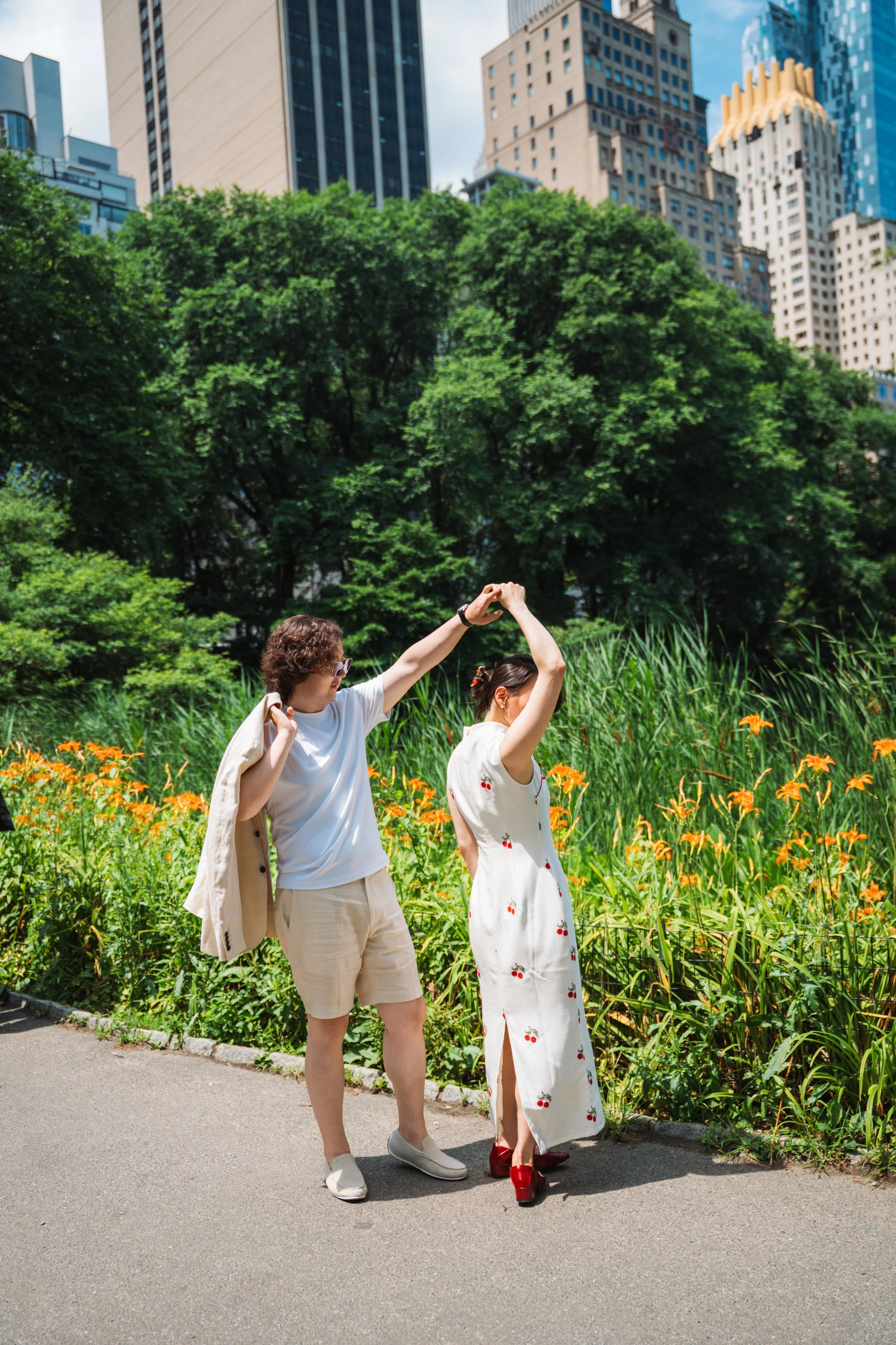 Two women are dancing in a park with tall green trees and skyscrapers in the background. One woman has curly hair and is wearing a white t-shirt and beige shorts, while the other is wearing a white dress with red cherries. They are holding hands and 