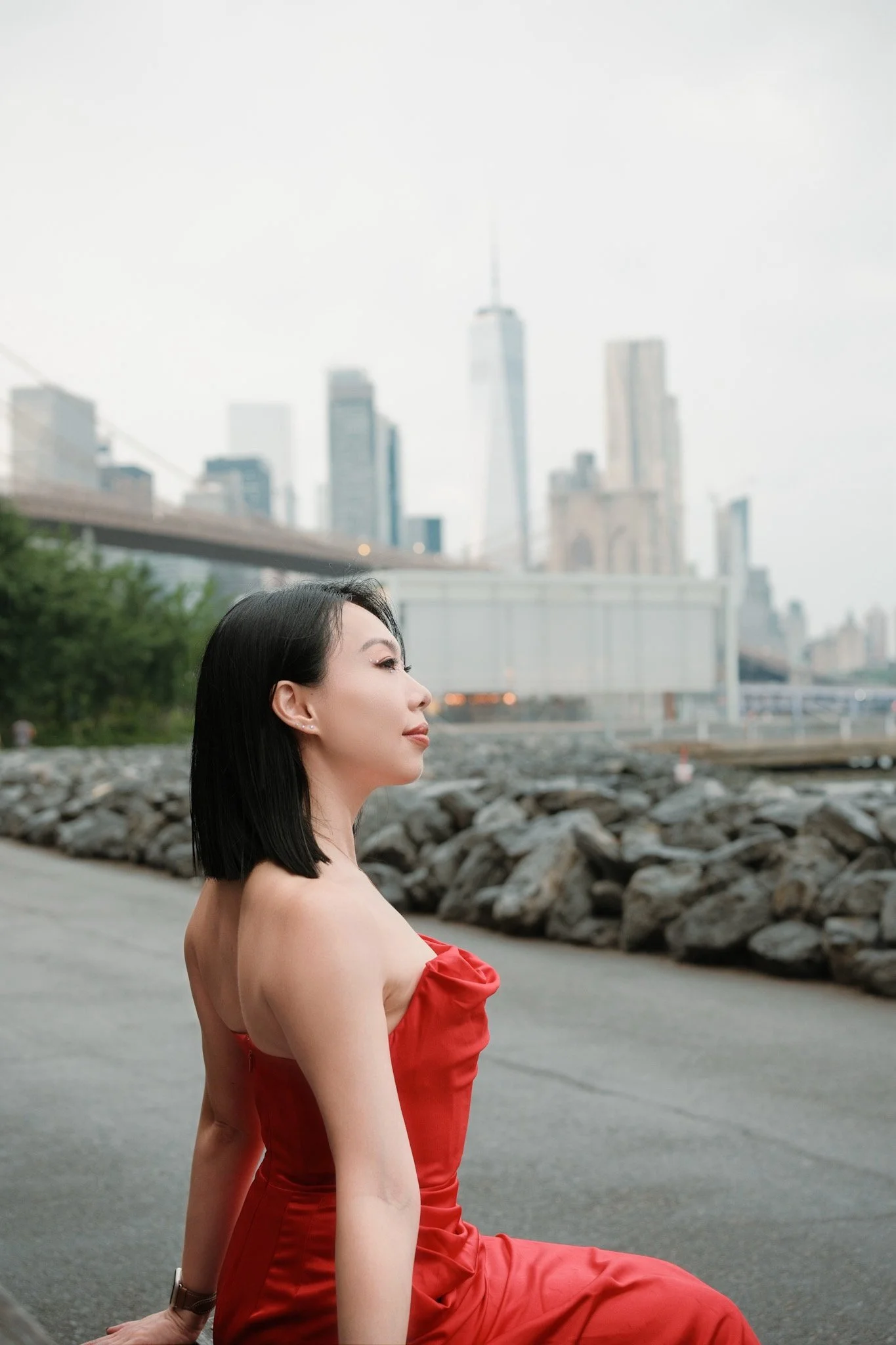 A woman with black hair wearing a red strapless dress, sitting outdoors on a rocky path with the New York City skyline in the background. The city skyscrapers, including the One World Trade Center, are blurred in the distance.