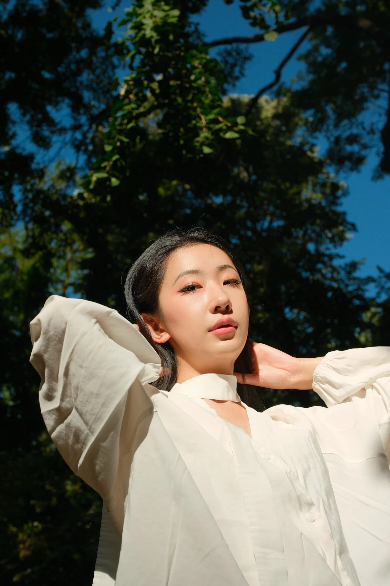 Portrait of a young woman with black hair, wearing a white jacket, outdoors with green trees and blue sky in the background.