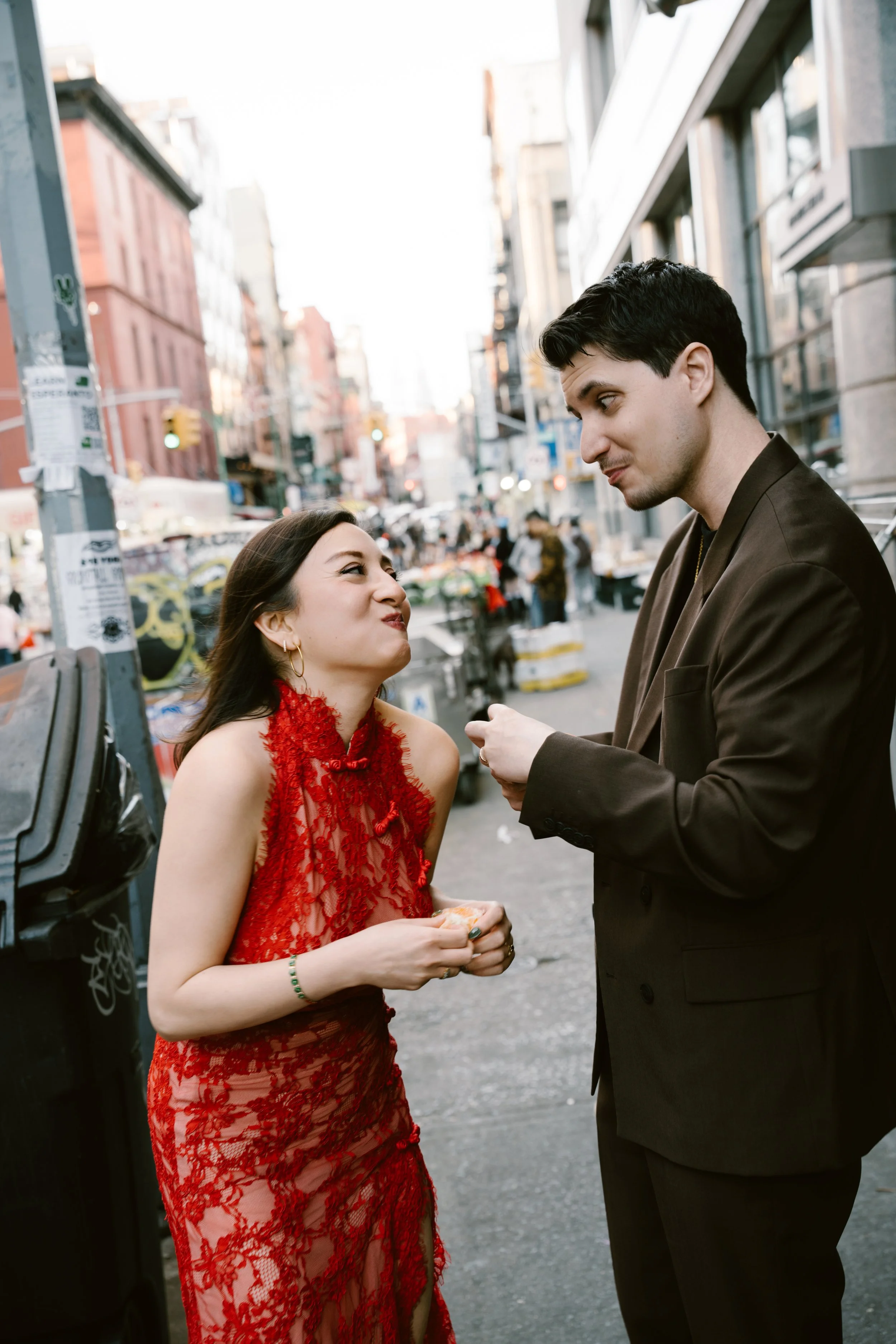 A man and woman are engaged in a conversation on a city street, with buildings and people in the background. The woman is wearing a red lace dress and the man is in a dark blazer.