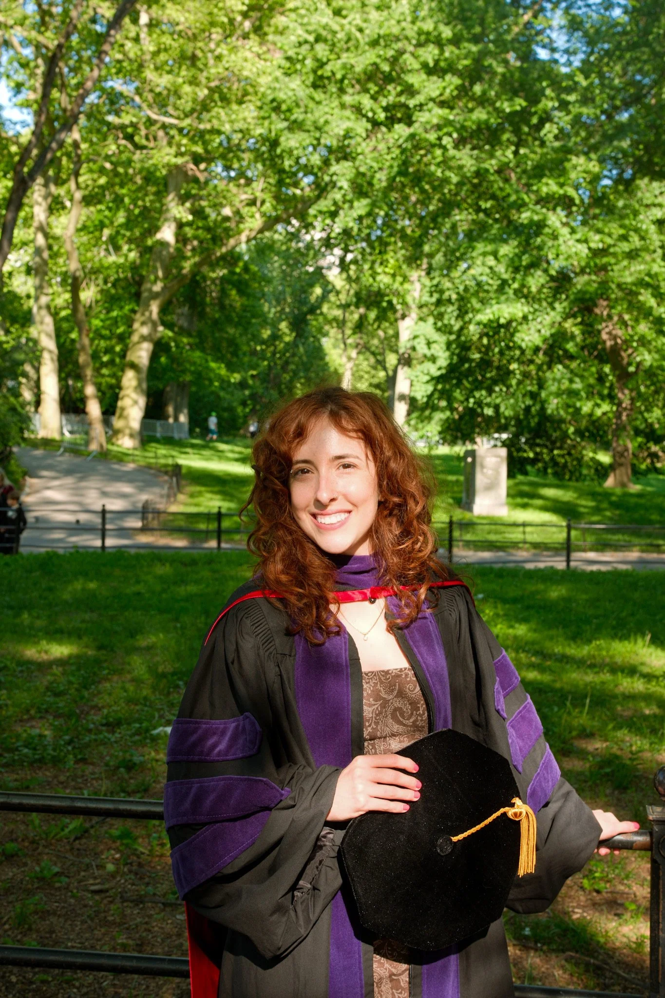 A woman with curly red hair smiling outdoors in a park, wearing a black graduation gown with purple and red accents, holding a black graduation cap with a gold tassel.