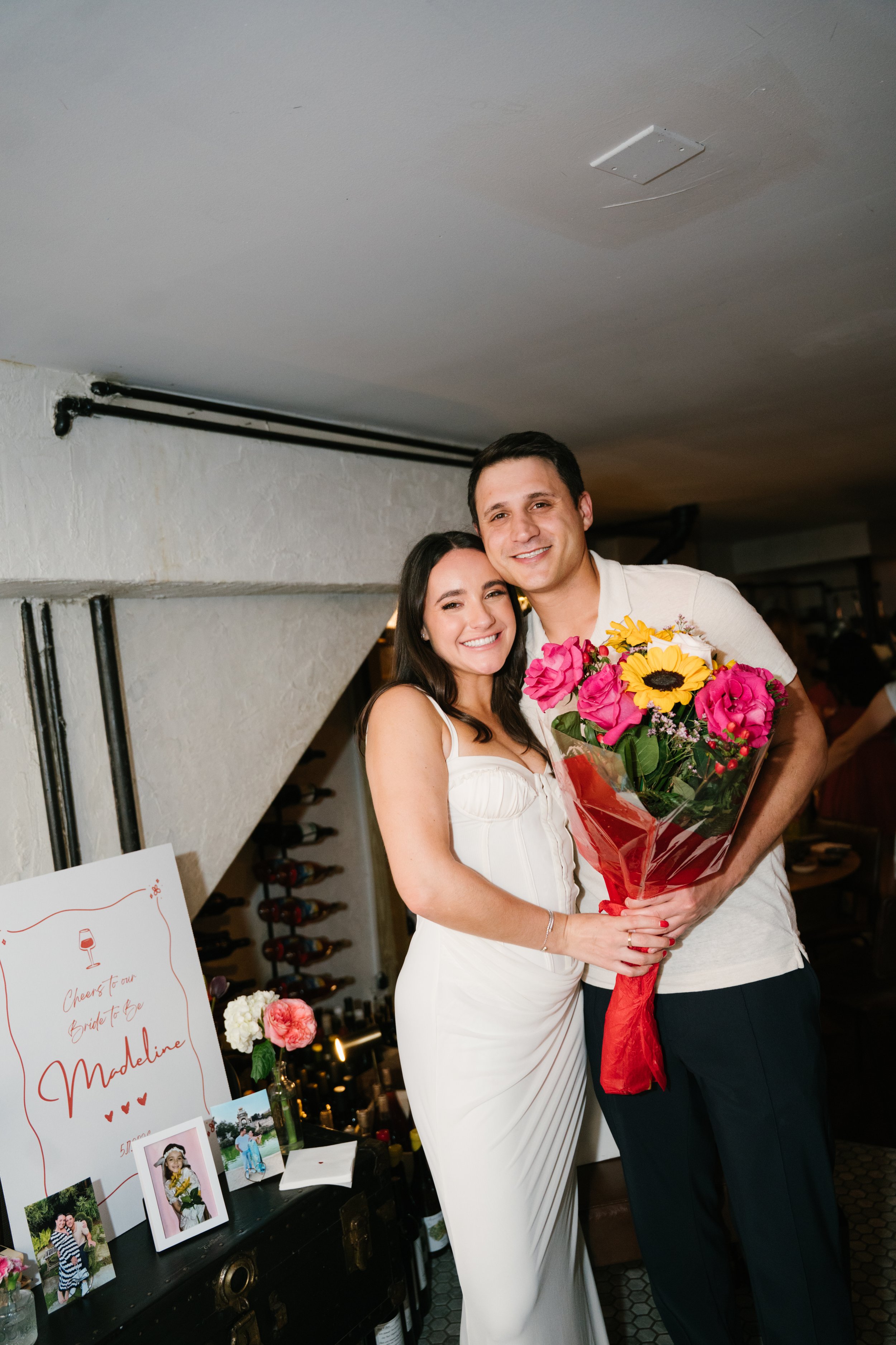 A happy couple, the woman in a white dress holding a bouquet of colorful flowers and the man in a light shirt, standing close together and smiling at an indoor celebration, possibly a wedding.