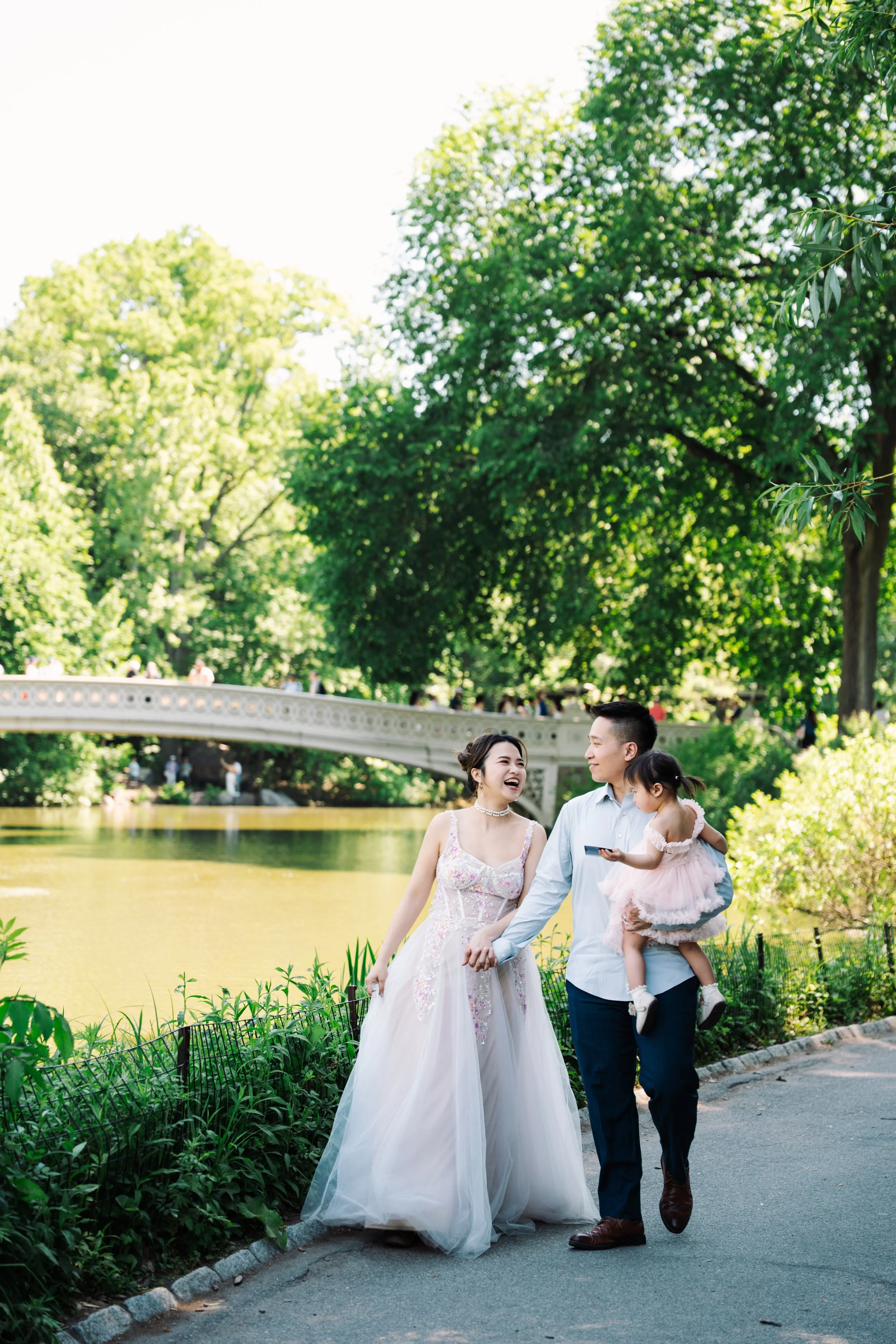 A happy couple dressed in wedding attire walking through a park by a lake, holding a young girl in a pink dress, with lush green trees and a bridge in the background.