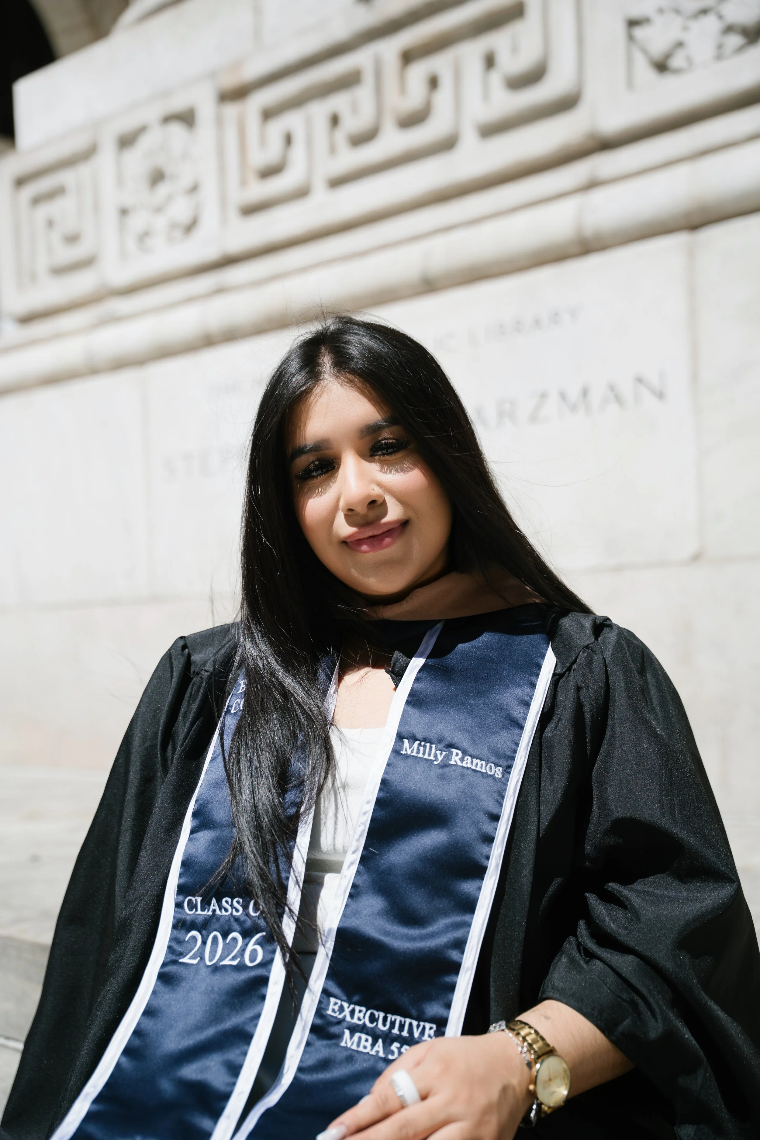 Young woman with long dark hair wearing a graduation gown and cap, standing outside in front of a white stone wall, smiling at the camera. She has a watch and ring on her left hand and a sash with her name and degree.