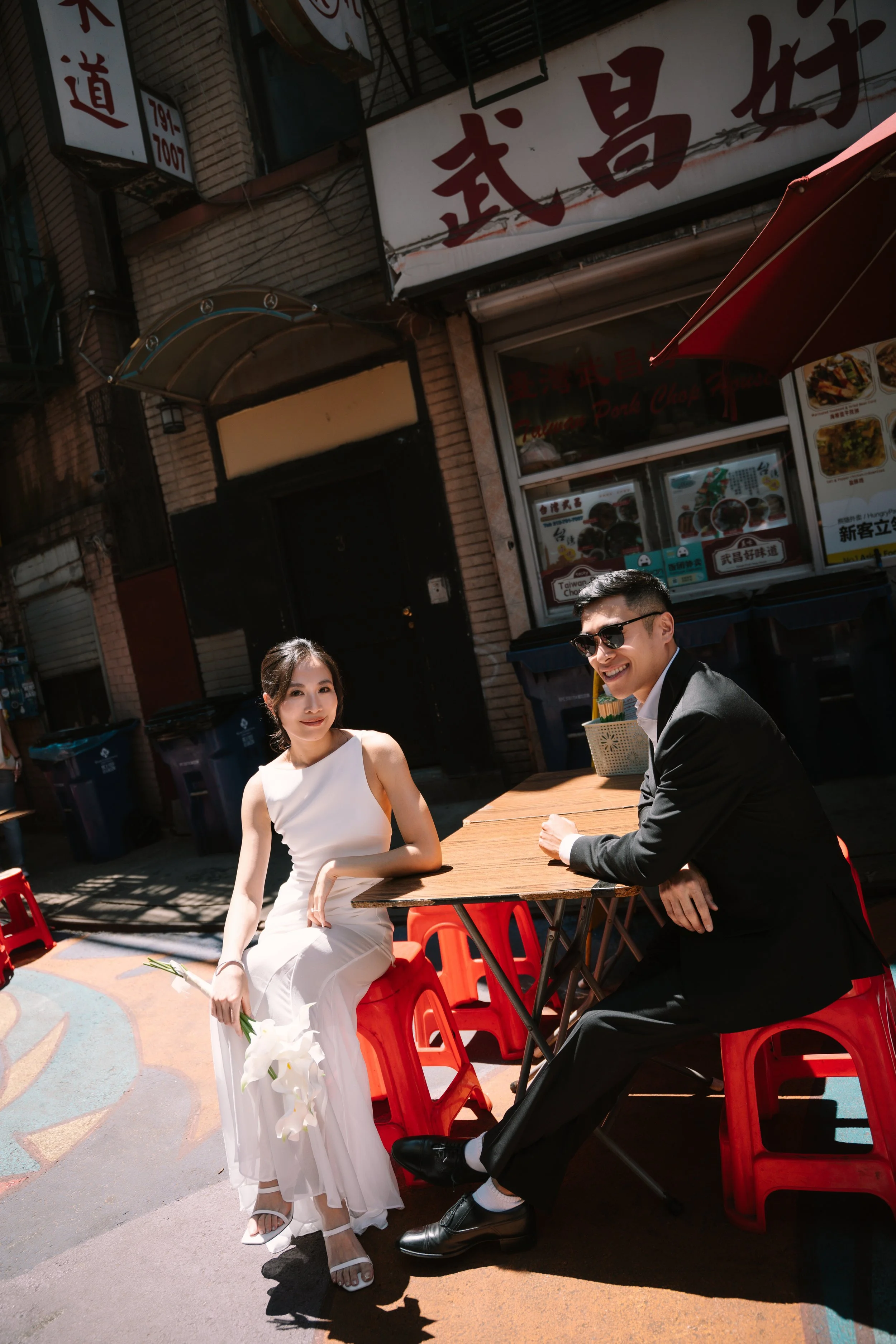 A smiling woman in a white dress holding white calla lilies beside a man in a suit sitting at a wooden outdoor table in an urban street scene.