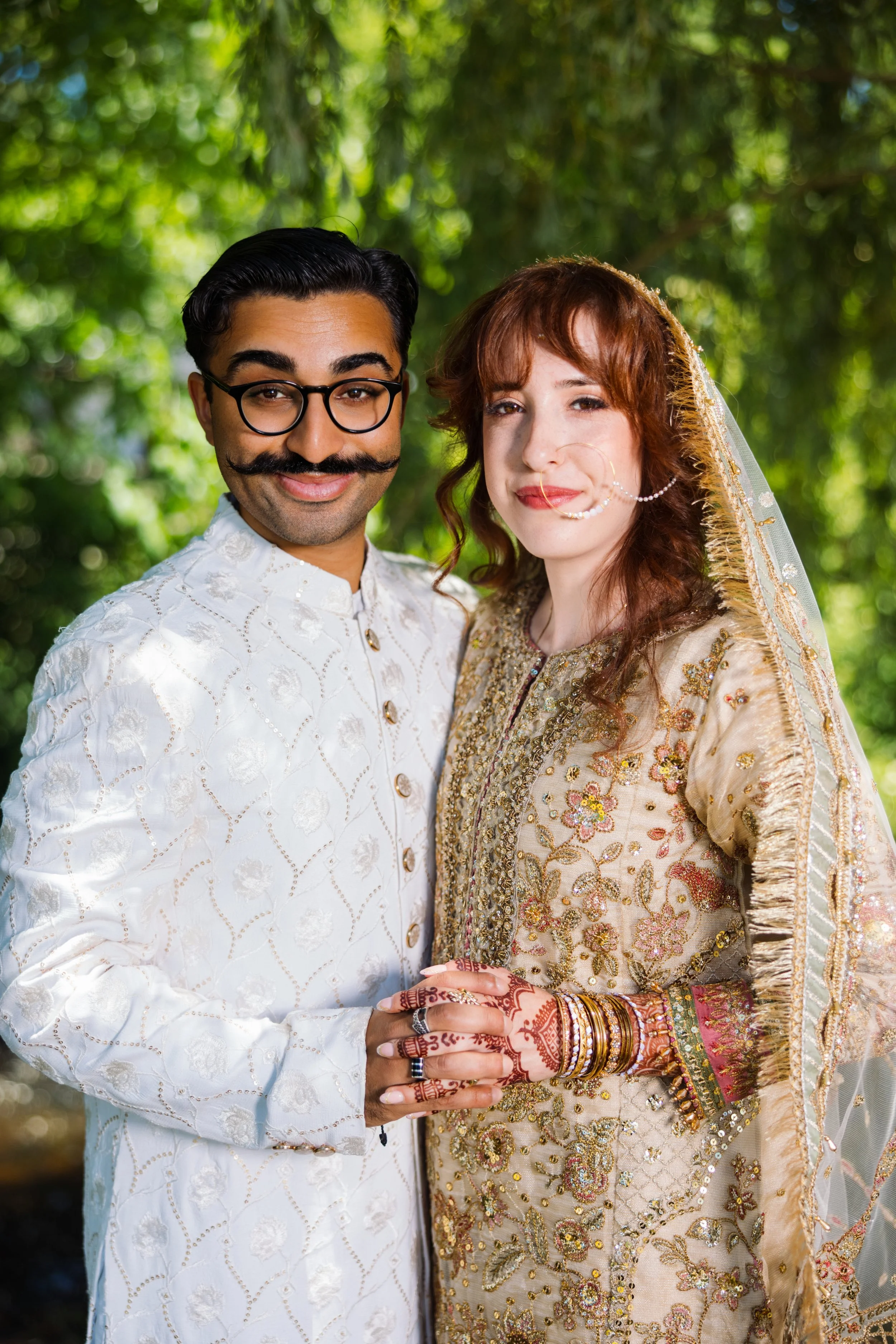 A man and a woman in traditional Indian attire standing outdoors with green foliage in the background, holding hands and smiling.