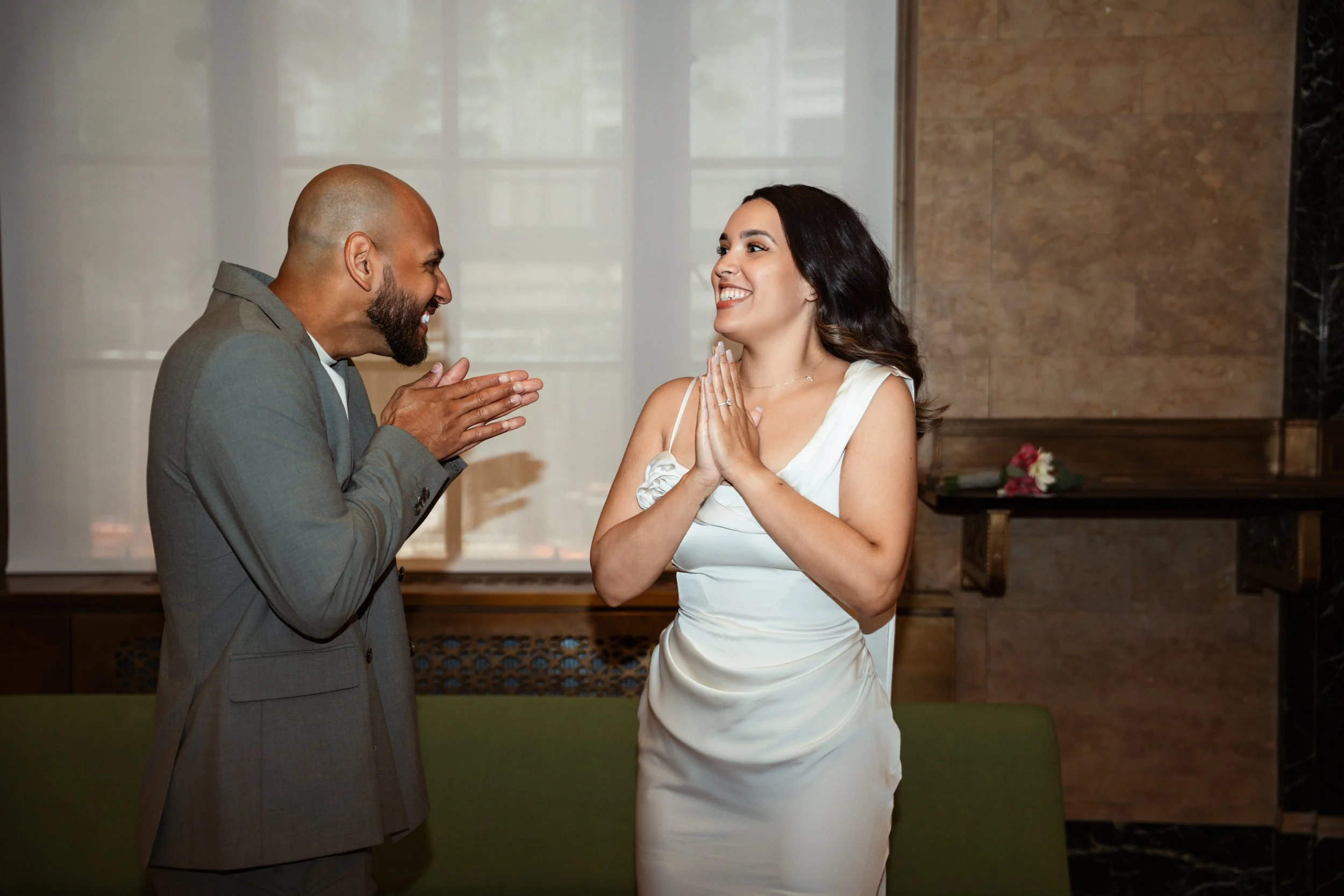A man and woman are facing each other with smiles; the man has his hands together as if praying or pleading, and the woman has her hands together near her chest, both are dressed formally and are indoors.