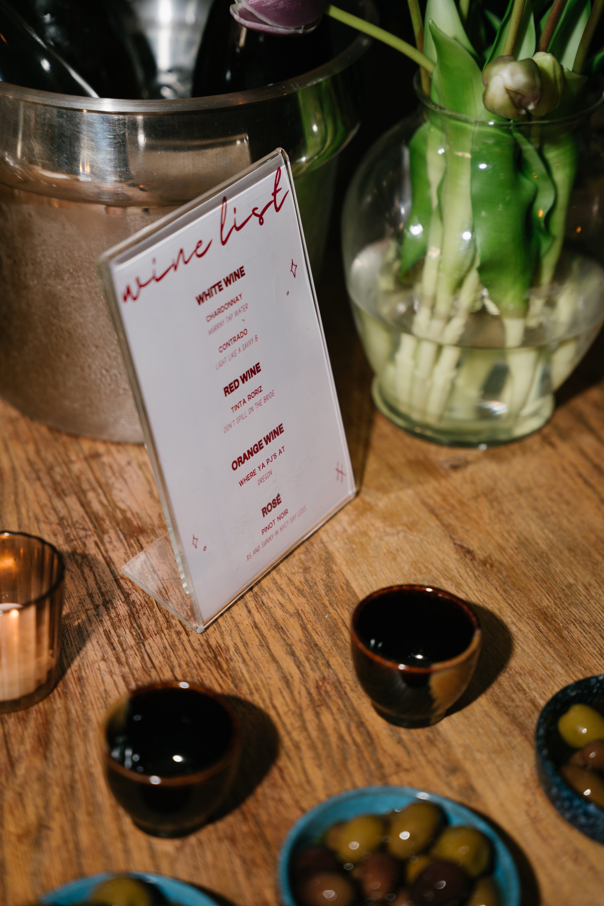 A wooden table with small bowls containing various types of olives, a glass vase with green plants, and a menu sign listing wine options.