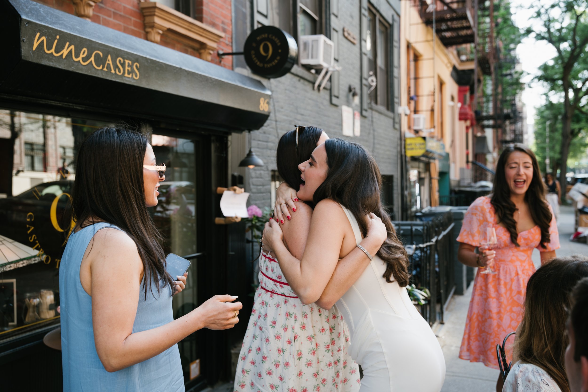 Three women are outside on a city sidewalk, hugging and smiling. One woman is holding a glass of wine, and another is holding a cell phone. They are standing next to a storefront with a sign that reads 'nine cases' and have various street-facing hous