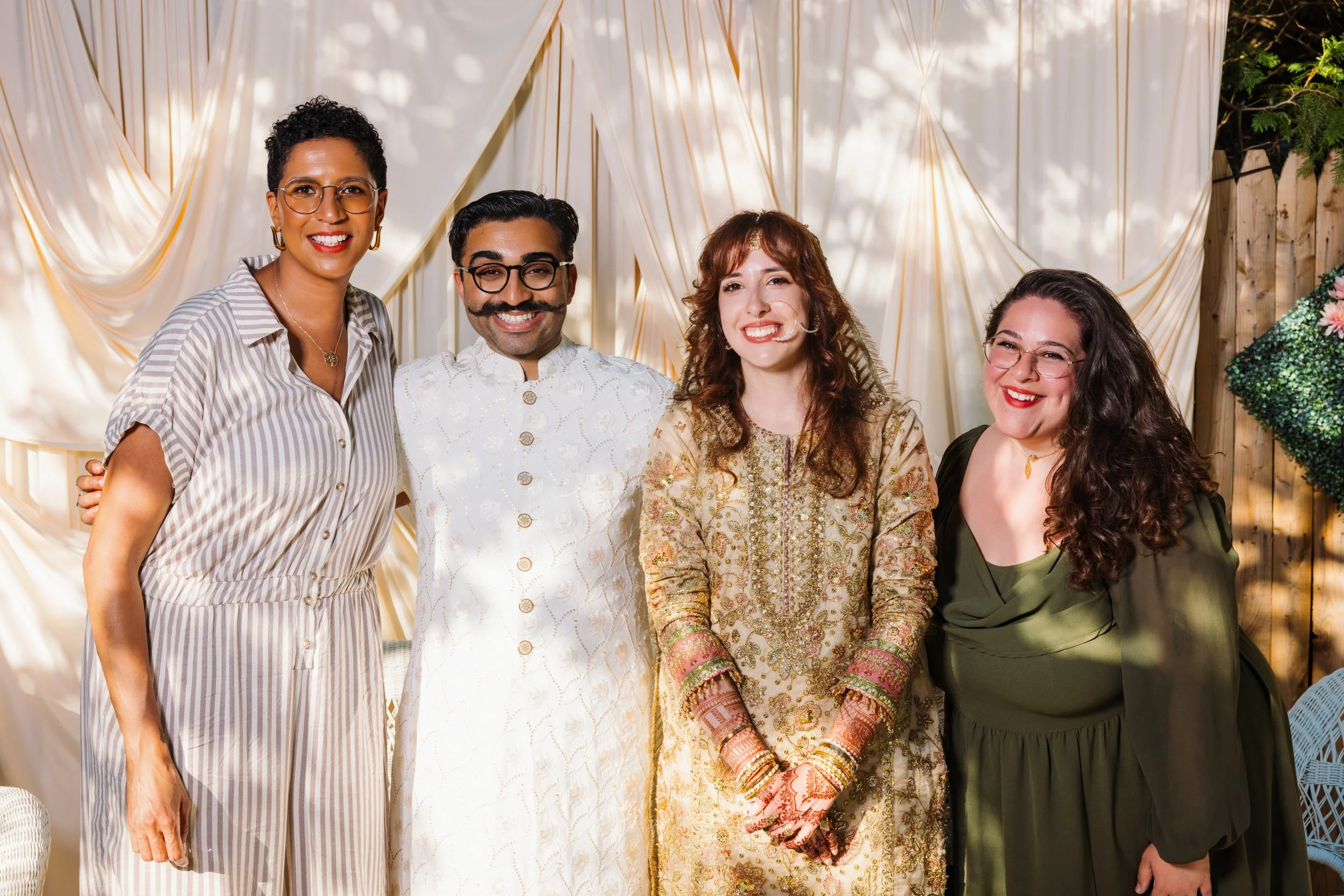Group of five diverse women smiling at a celebration or gathering, standing in front of a white draped backdrop and a wooden fence, dressed in various outfits.