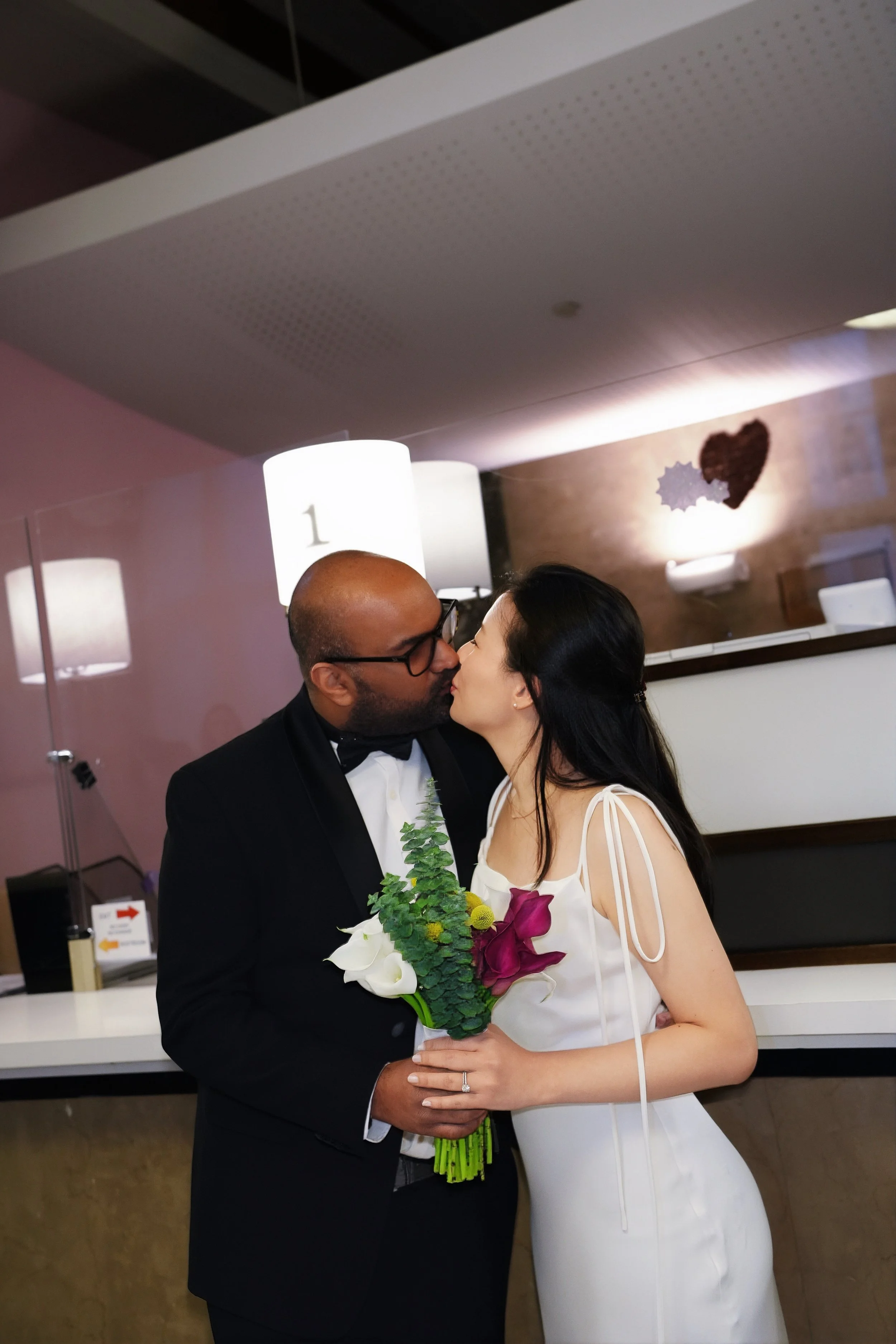 A couple kissing at a wedding or celebration, the man wearing a tuxedo and the woman wearing a white dress holding a bouquet of flowers.