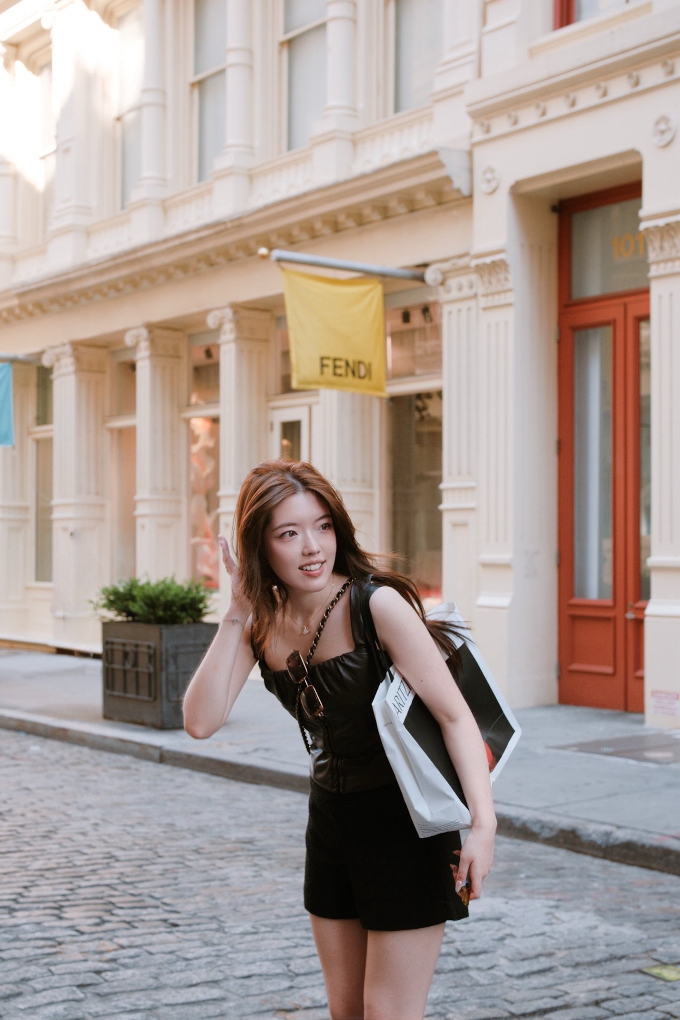 A young woman with brown hair in a black dress stands on a cobblestone street in front of a building with white ornate architecture and a yellow "FENDI" banner. She is holding a shopping bag and a smartphone, smiling slightly and adjusting her hair.