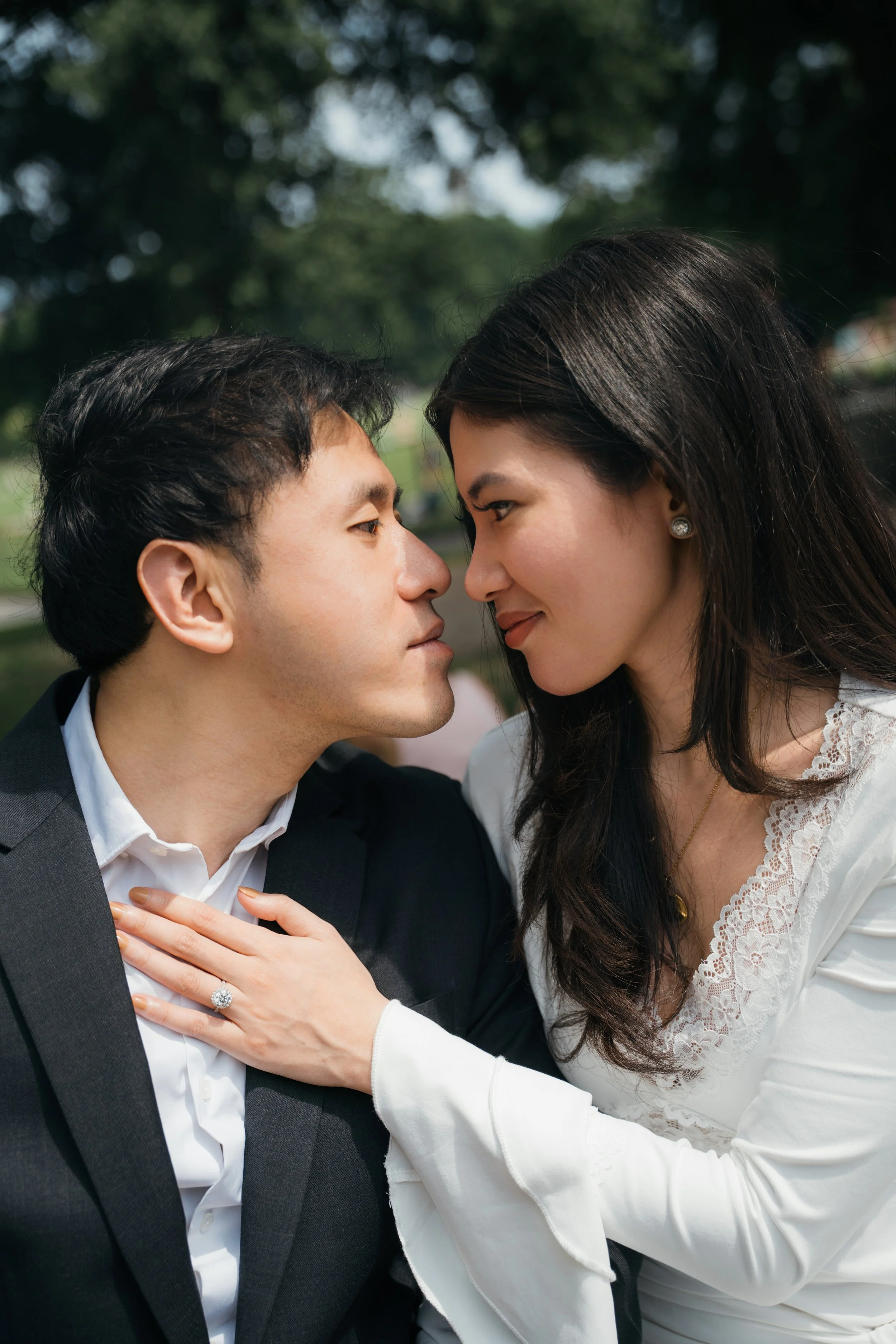 A couple is close, touching foreheads and smiling, outdoors with trees in the background.
