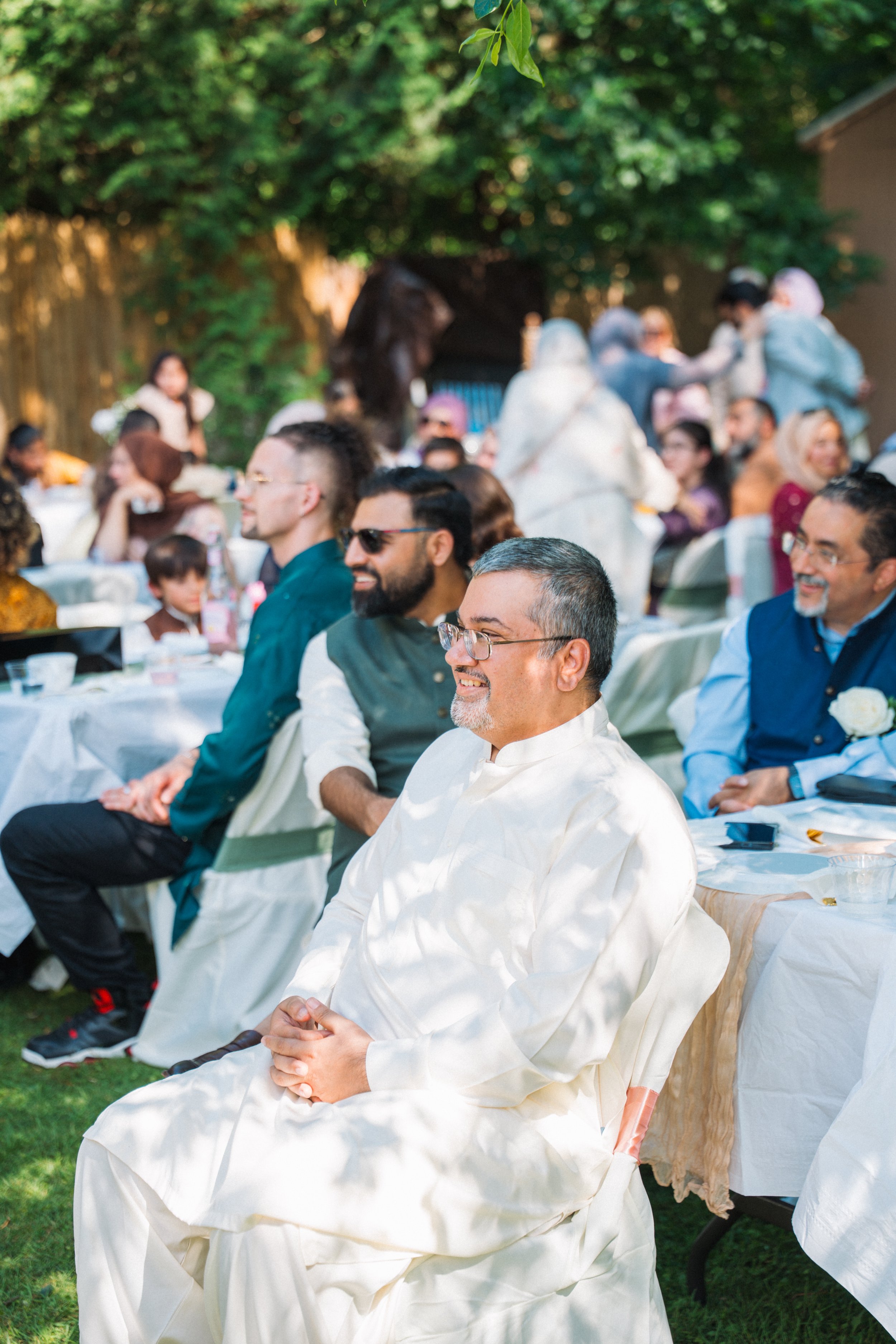 People gathered at an outdoor event, sitting at tables, with some standing and talking under a tree in a backyard setting.