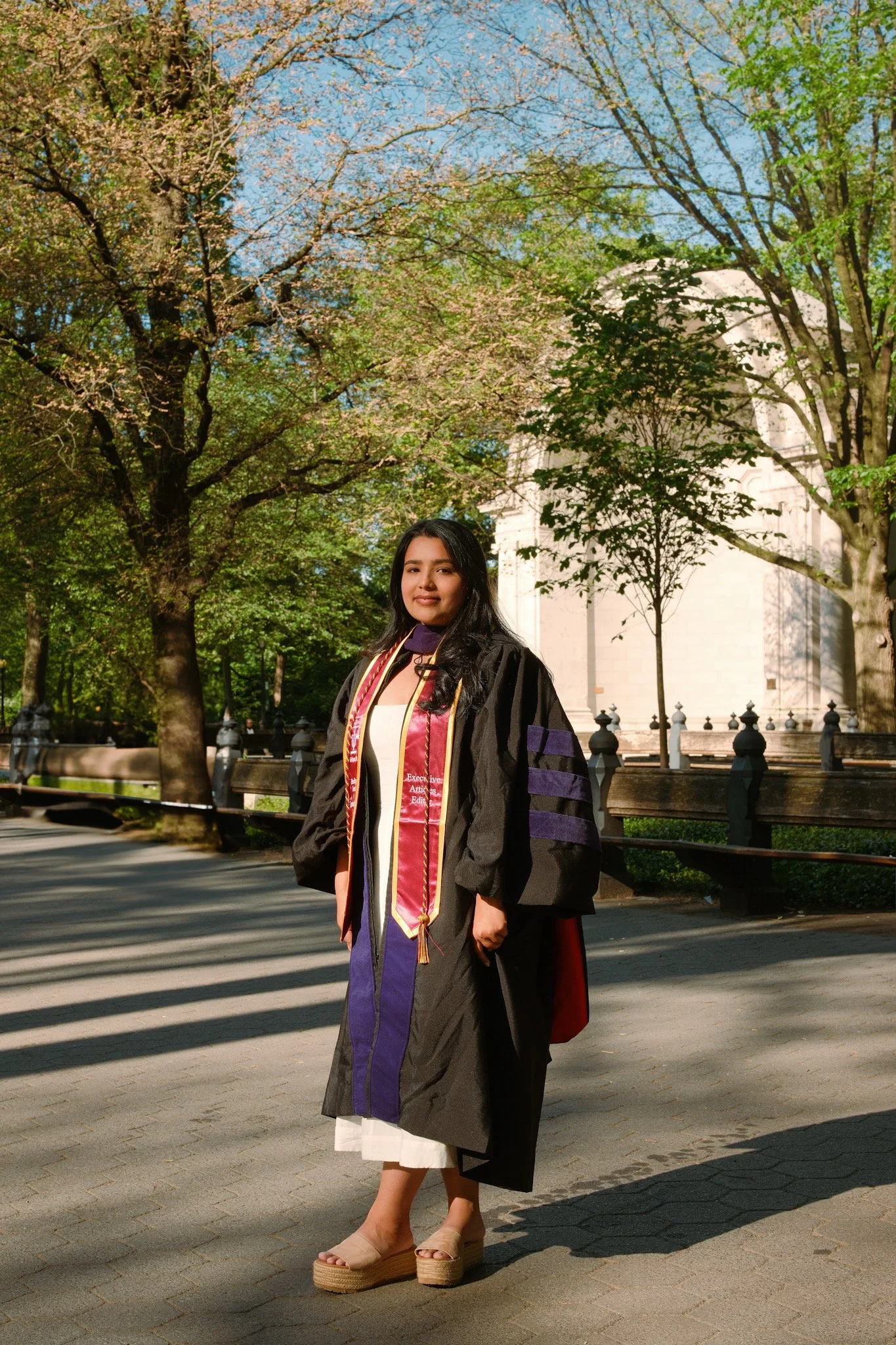 A young woman in graduation attire standing outdoors on a sunny day with trees and a historic building in the background. She is wearing a black gown with purple accents, a white dress underneath, beige platform shoes, and multiple honor cords around