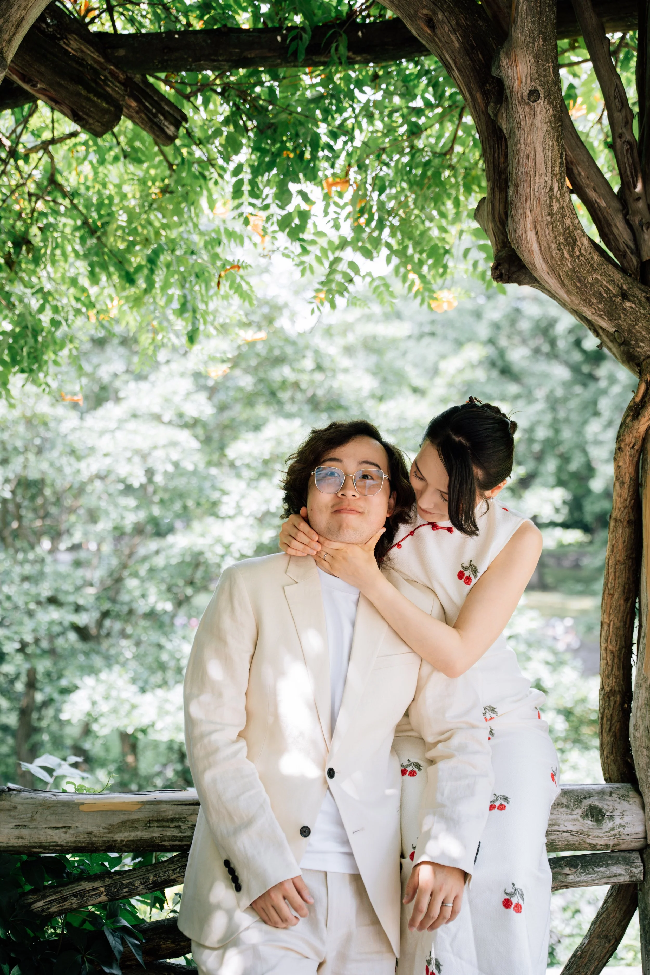 A couple dressed in light-colored clothing standing outdoors beneath a large tree with green leaves.