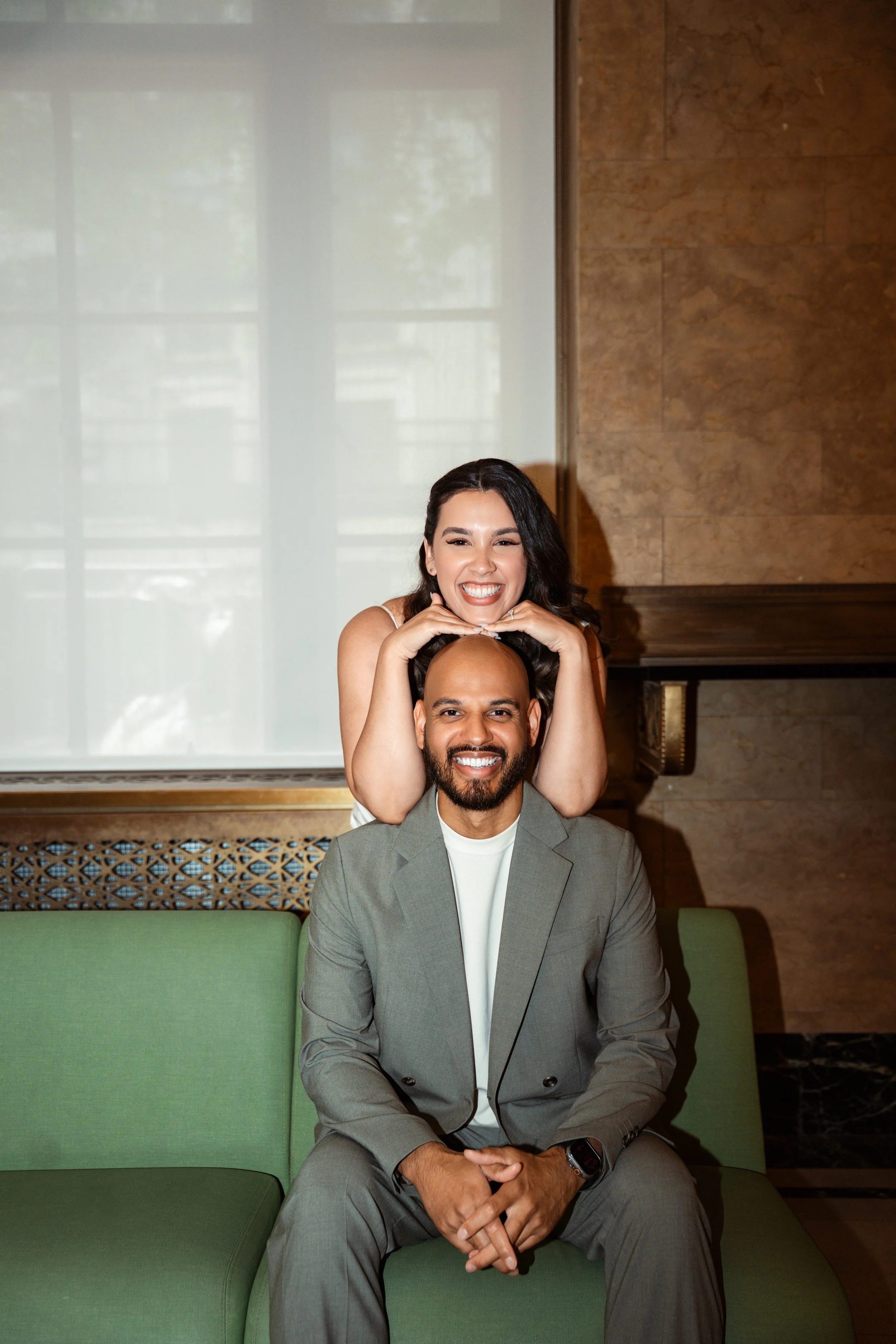 A man sitting on a green sofa with a woman standing behind him, smiling and resting her chin on his head in a luxurious room with a marble wall and large window.