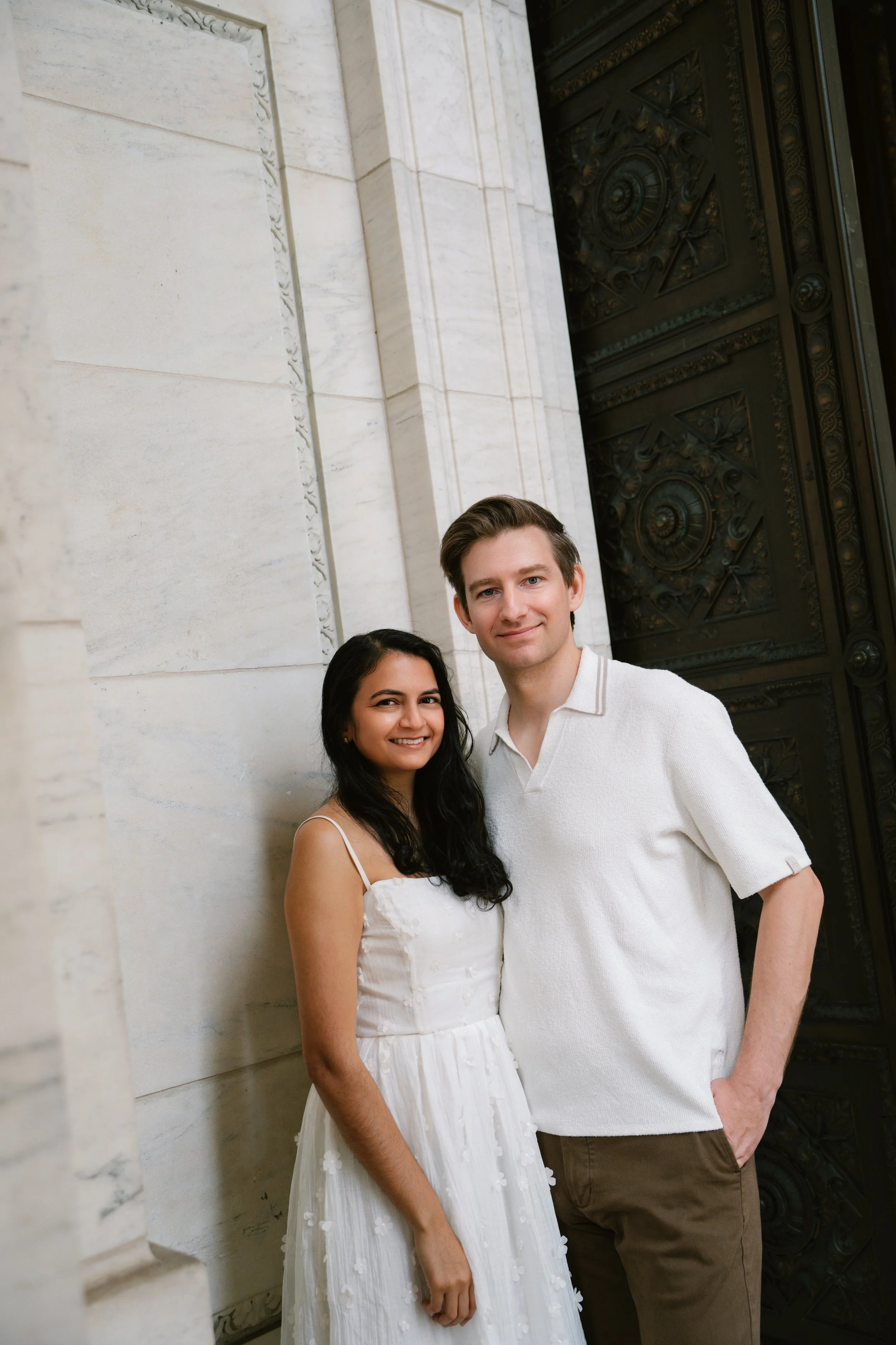 A young couple standing together outdoors in front of a large decorative metal door and a white stone wall. The woman is wearing a white dress with floral appliqués, and the man is wearing a white polo shirt and brown pants. They are smiling and posi