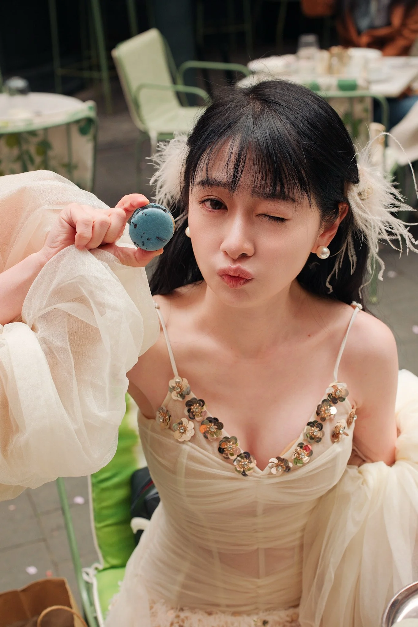A woman with dark hair, wearing pearl earrings and a beige dress decorated with floral embellishments, winks and makes a pouty face while holding a blue macaron.