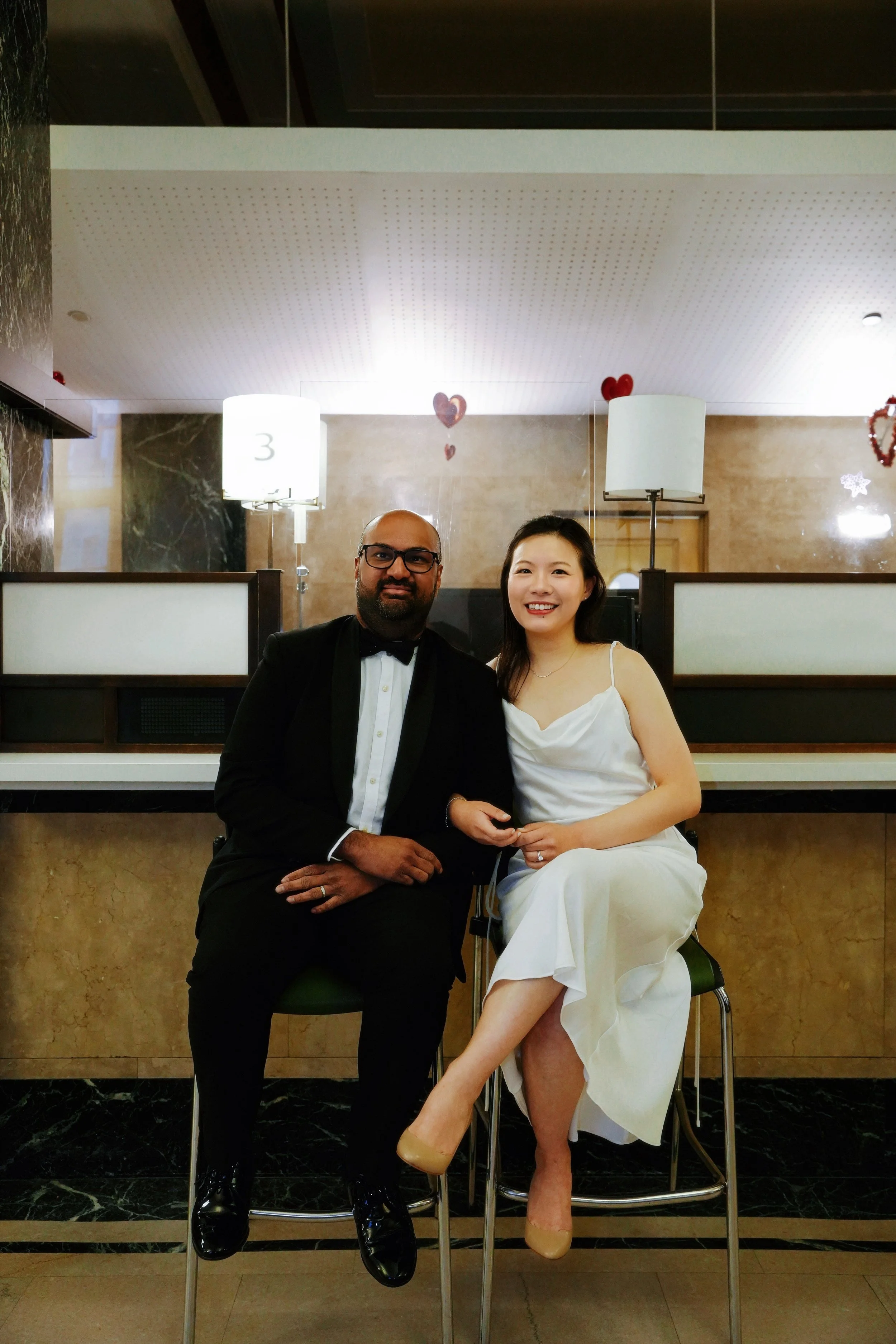 A man in a black tuxedo and a woman in a white dress sitting together in a restaurant, smiling at the camera, with Valentine's Day decorations including hearts in the background.