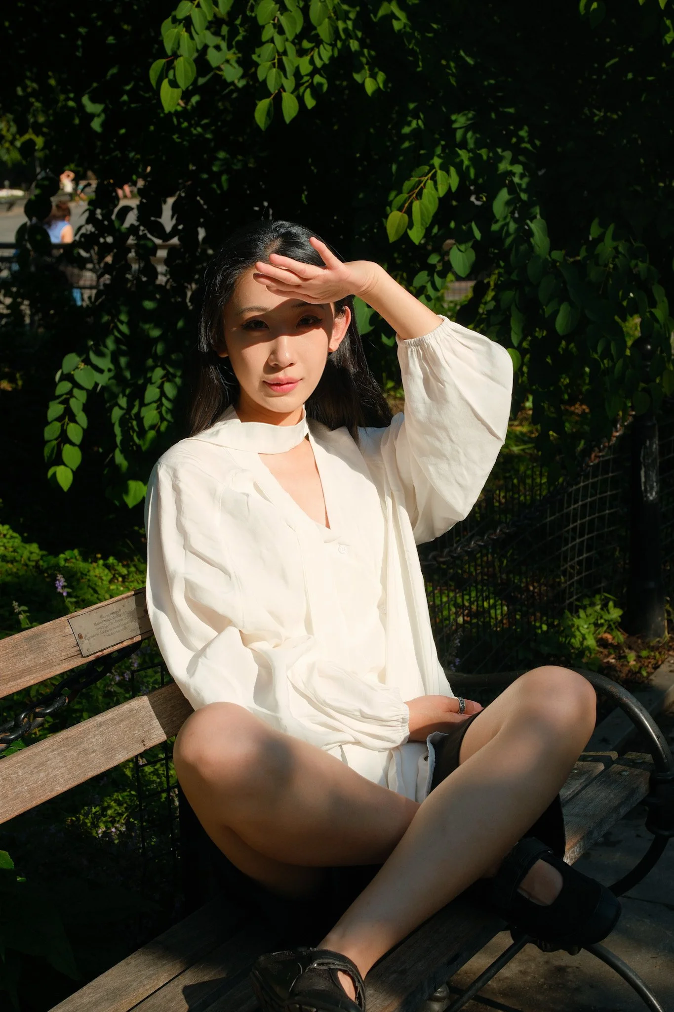 A young woman sitting on a park bench, shielding her eyes from the sun with her hand, wearing a white long-sleeve blouse, black shorts, and sandals, with green trees and sunlight in the background.