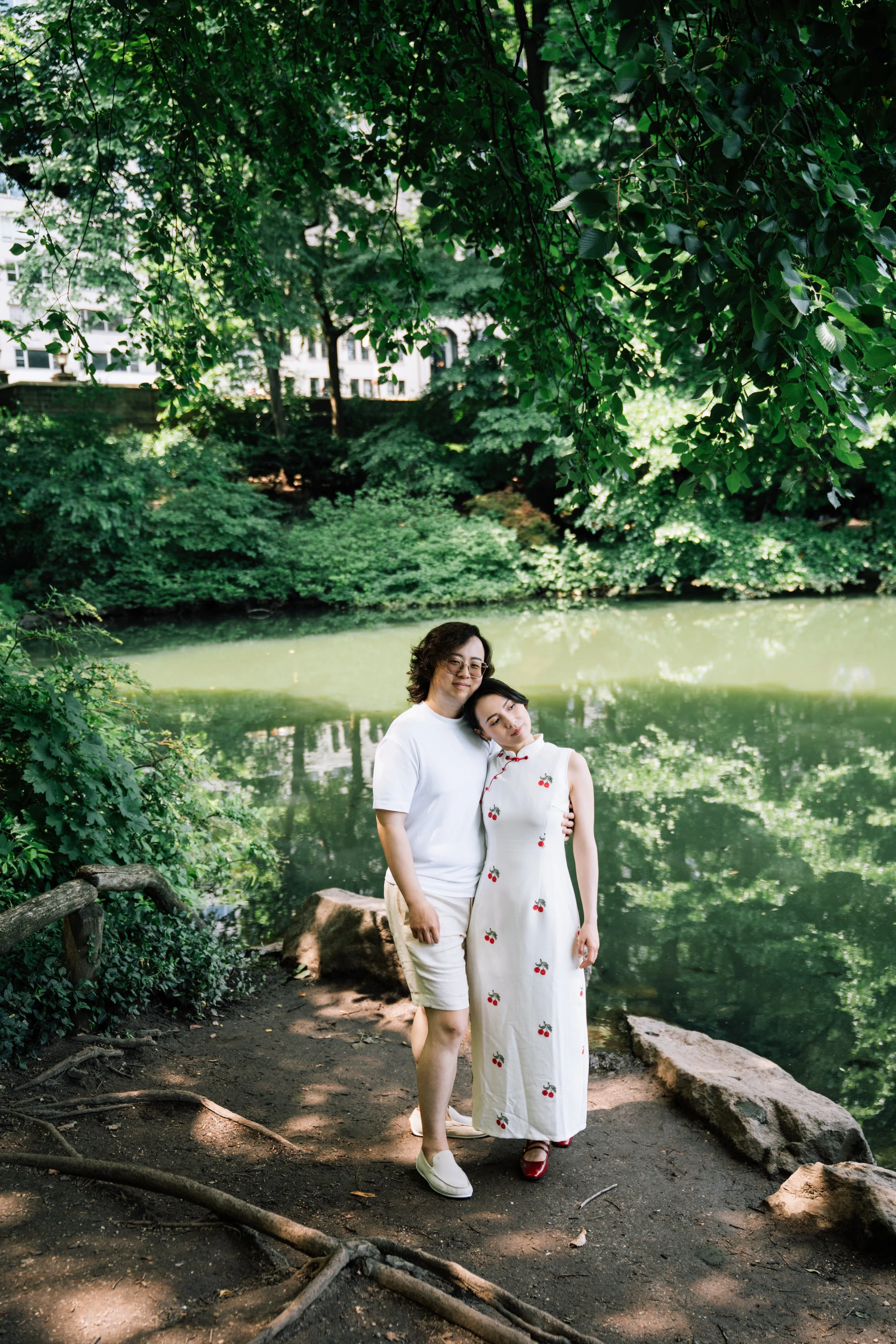 A couple stands together near a serene river in a lush, green park, surrounded by dense foliage and trees.