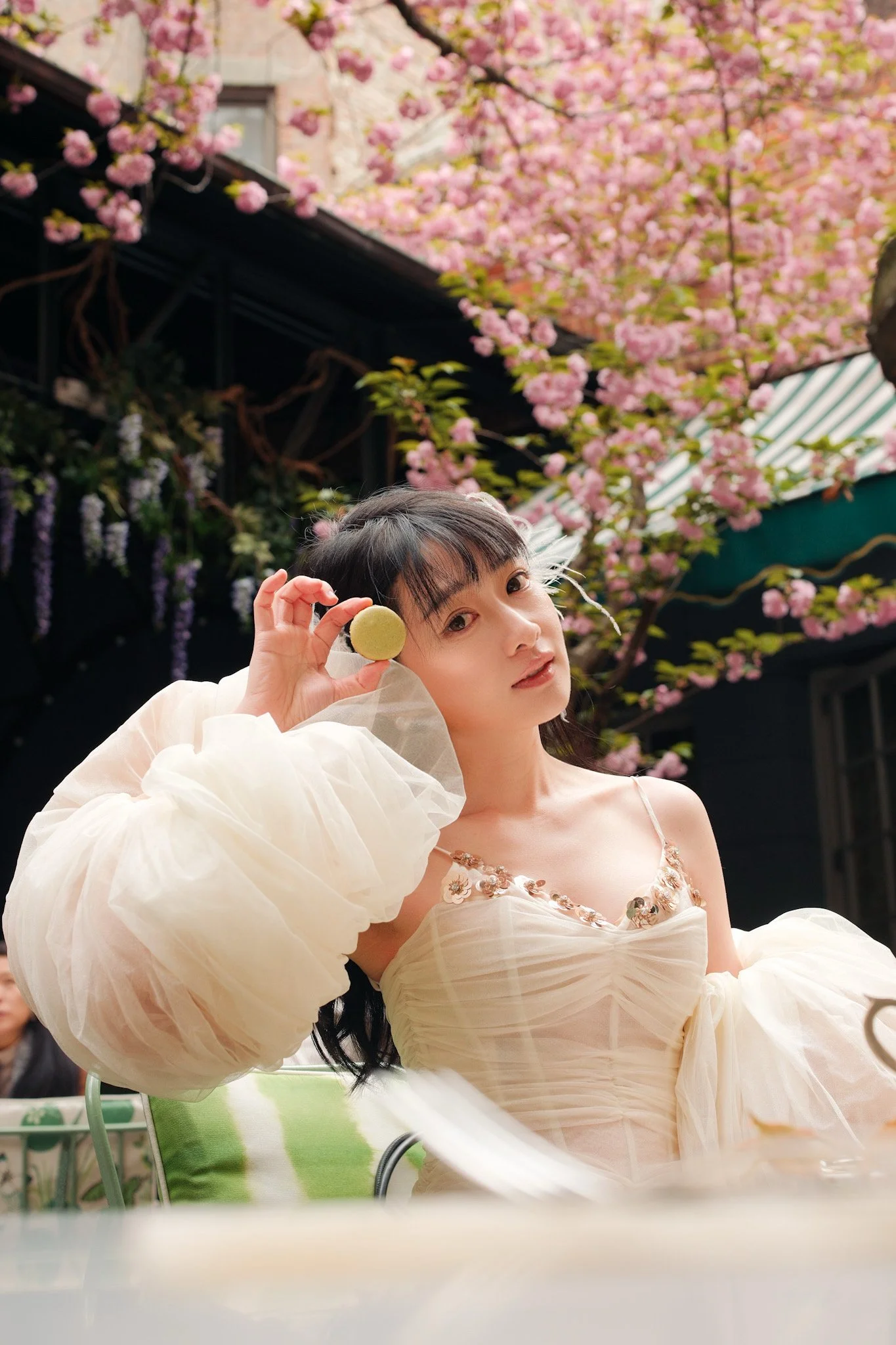 A woman with dark hair and fair skin holding a green macaroon near her face, wearing a beige dress with floral embroidery, with pink blossoming trees in the background.