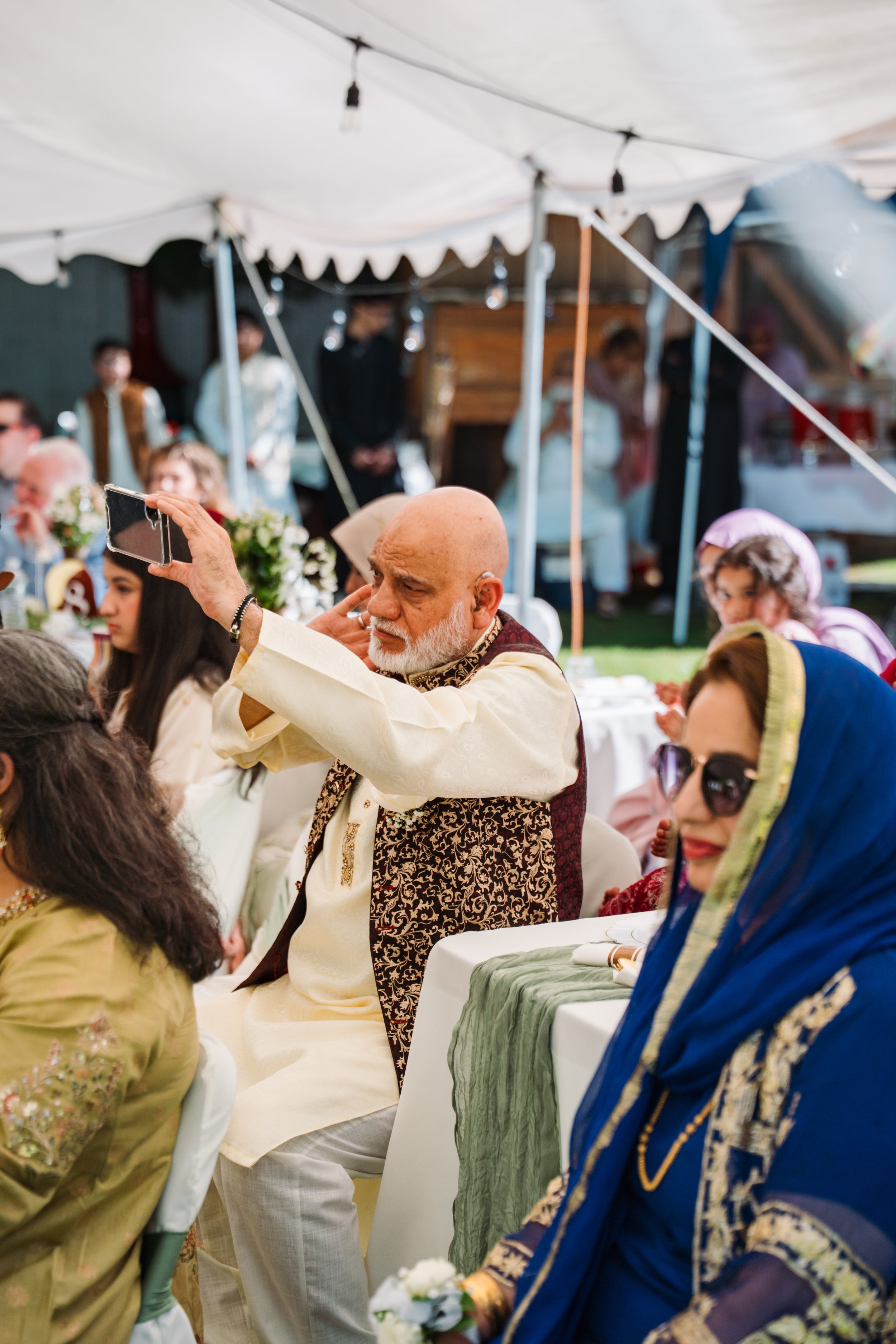 A group of people dressed in traditional South Asian clothing seated under a tent at a celebration, with some women wearing colorful sarees and men in kurta pajamas. One man in the foreground is taking a selfie with a smartphone.
