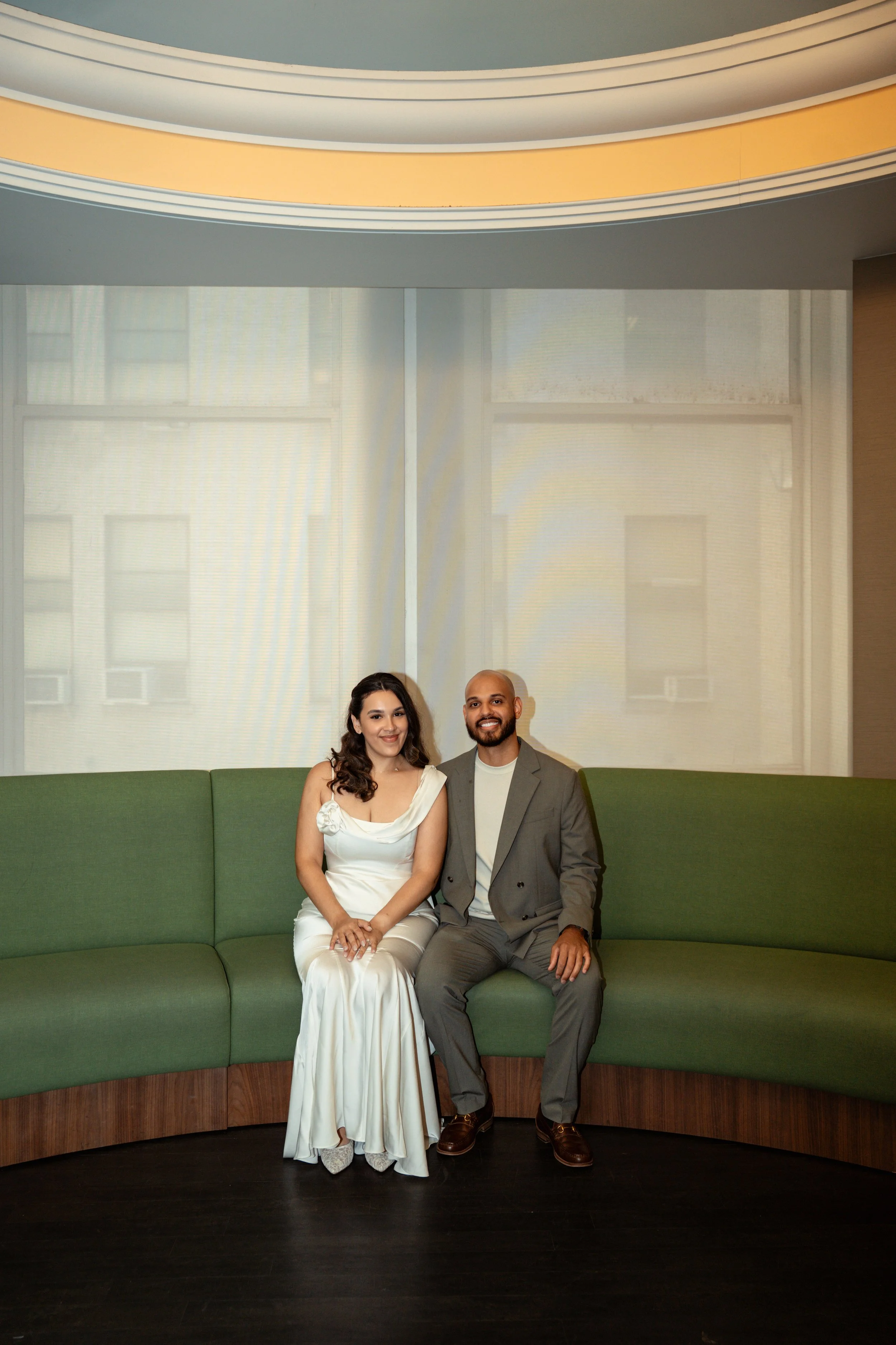 A man and a woman sitting on a green curved sofa in front of a window with sheer curtains, inside a room with gray walls and wooden flooring.