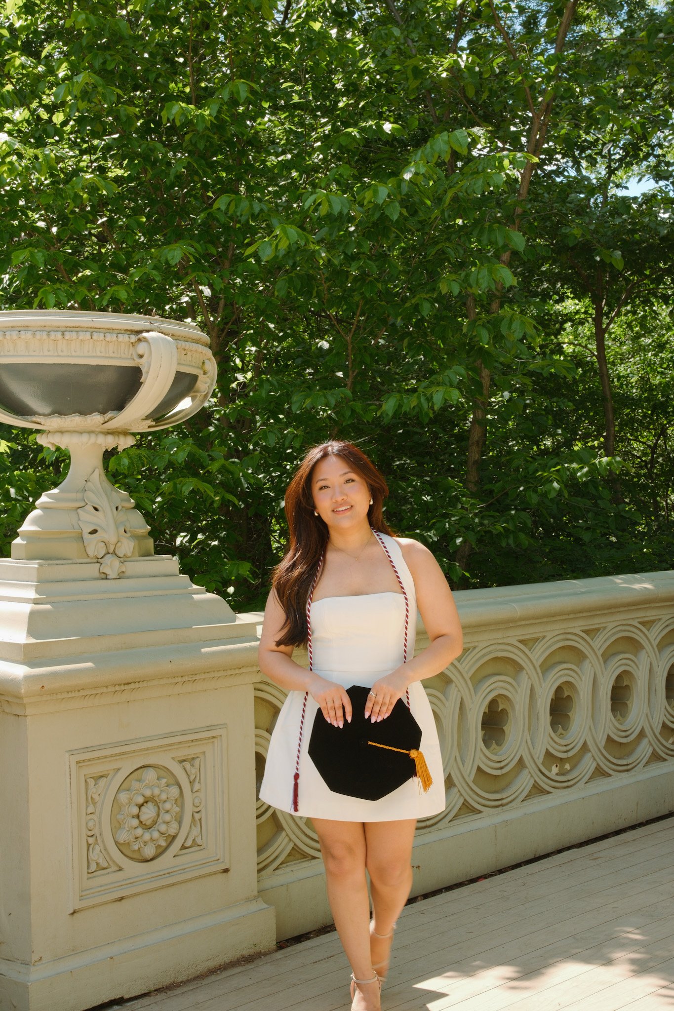 Young woman in a white dress holding a black graduation cap with an orange tassel, standing outdoors in front of a decorative rail and greenery.