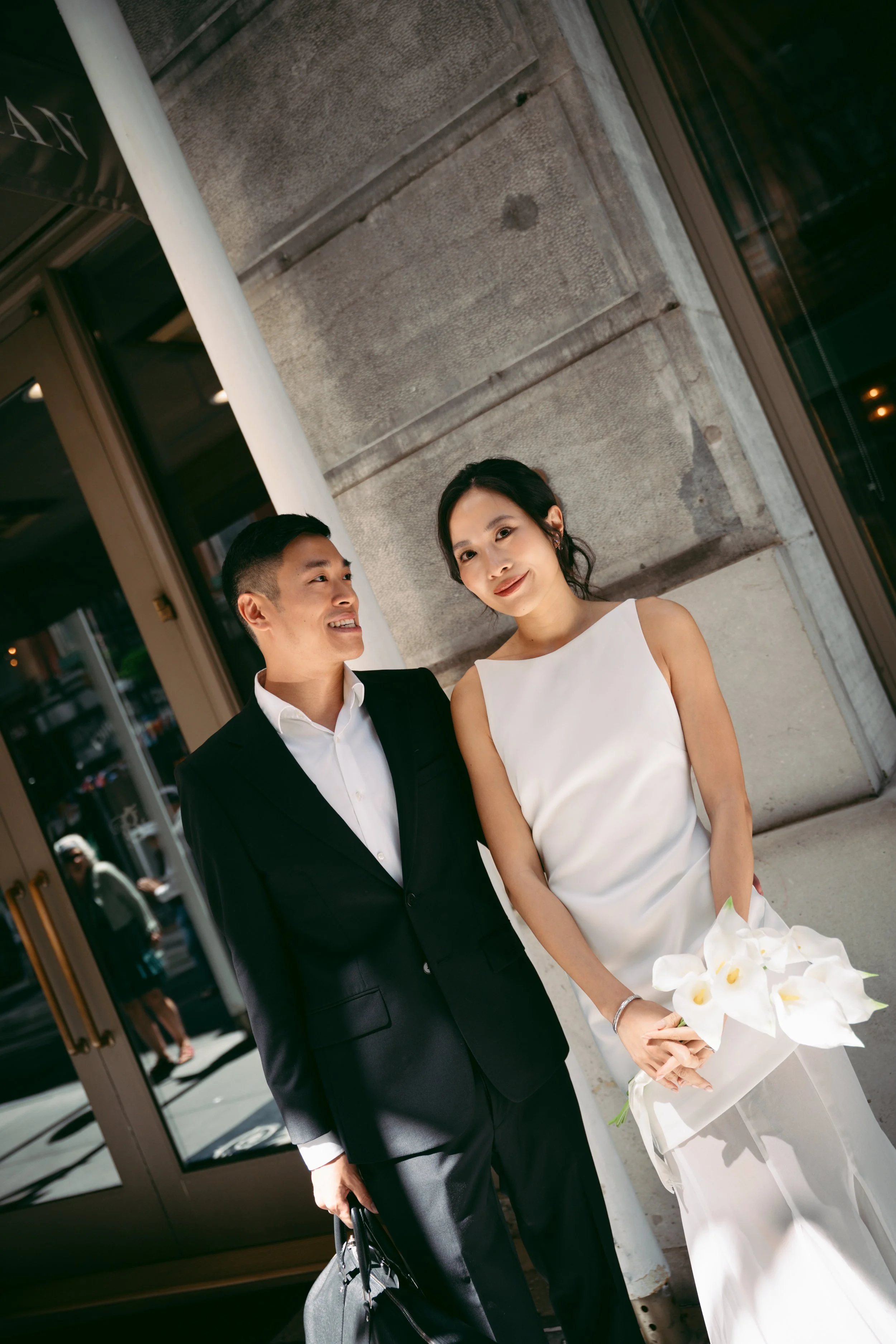 A man and woman, dressed formally, standing outside a building. The woman is holding a bouquet of white flowers. They are smiling and looking at each other.