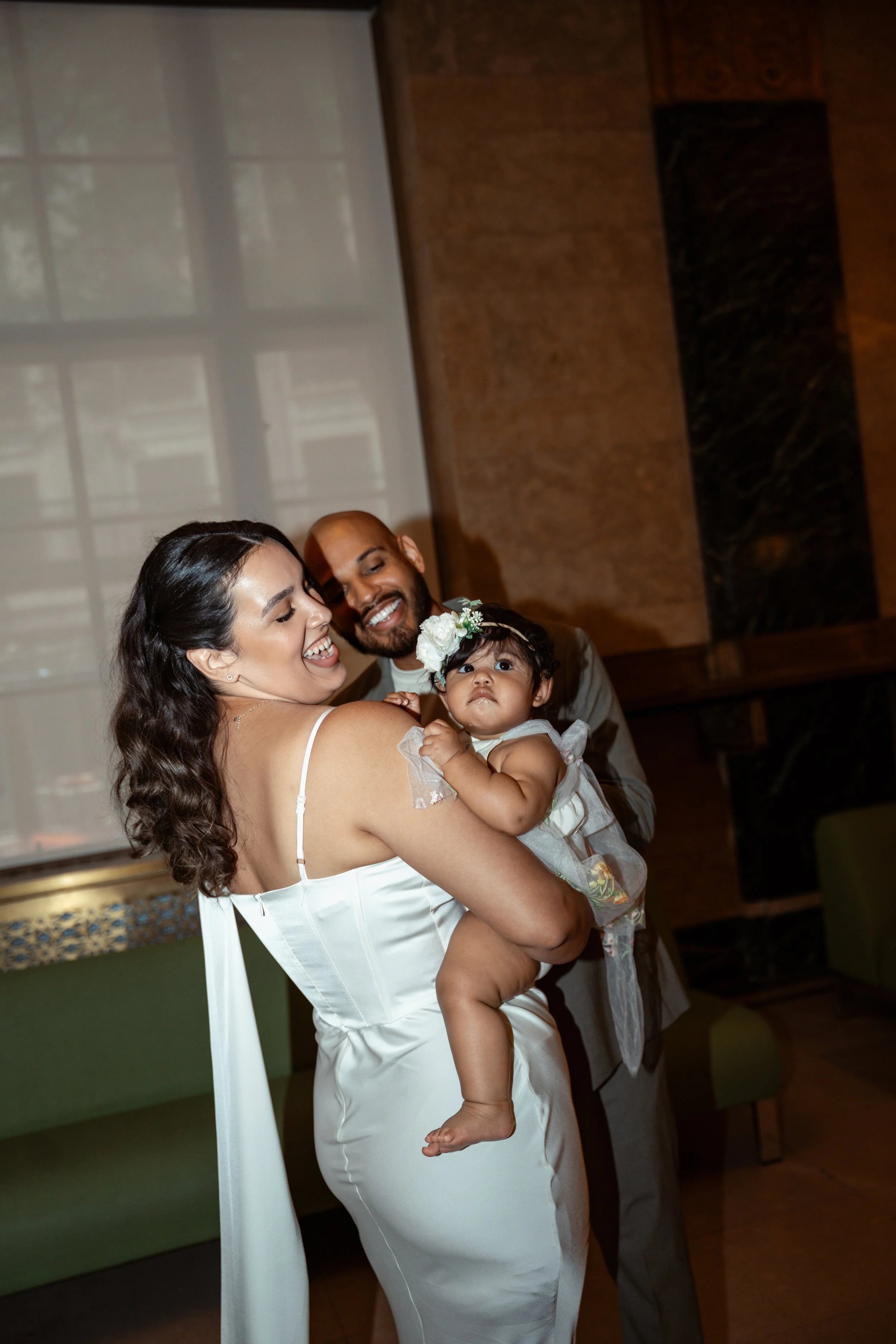 A woman in a white dress holding a young girl with a floral headband, with a man smiling behind them in an indoor setting.
