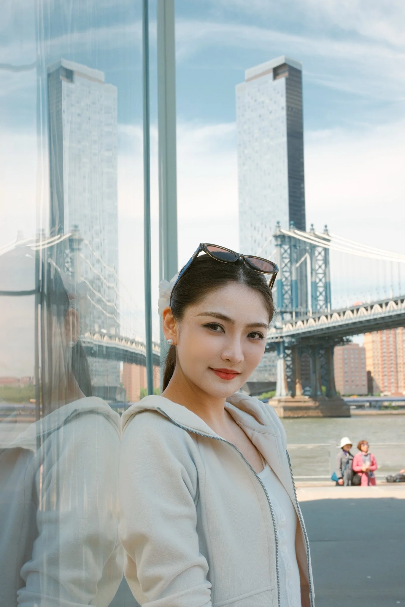 A young woman with sunglasses on her head, wearing a white zip-up hoodie, standing next to a reflective glass wall with the Brooklyn Bridge and city buildings in the background.